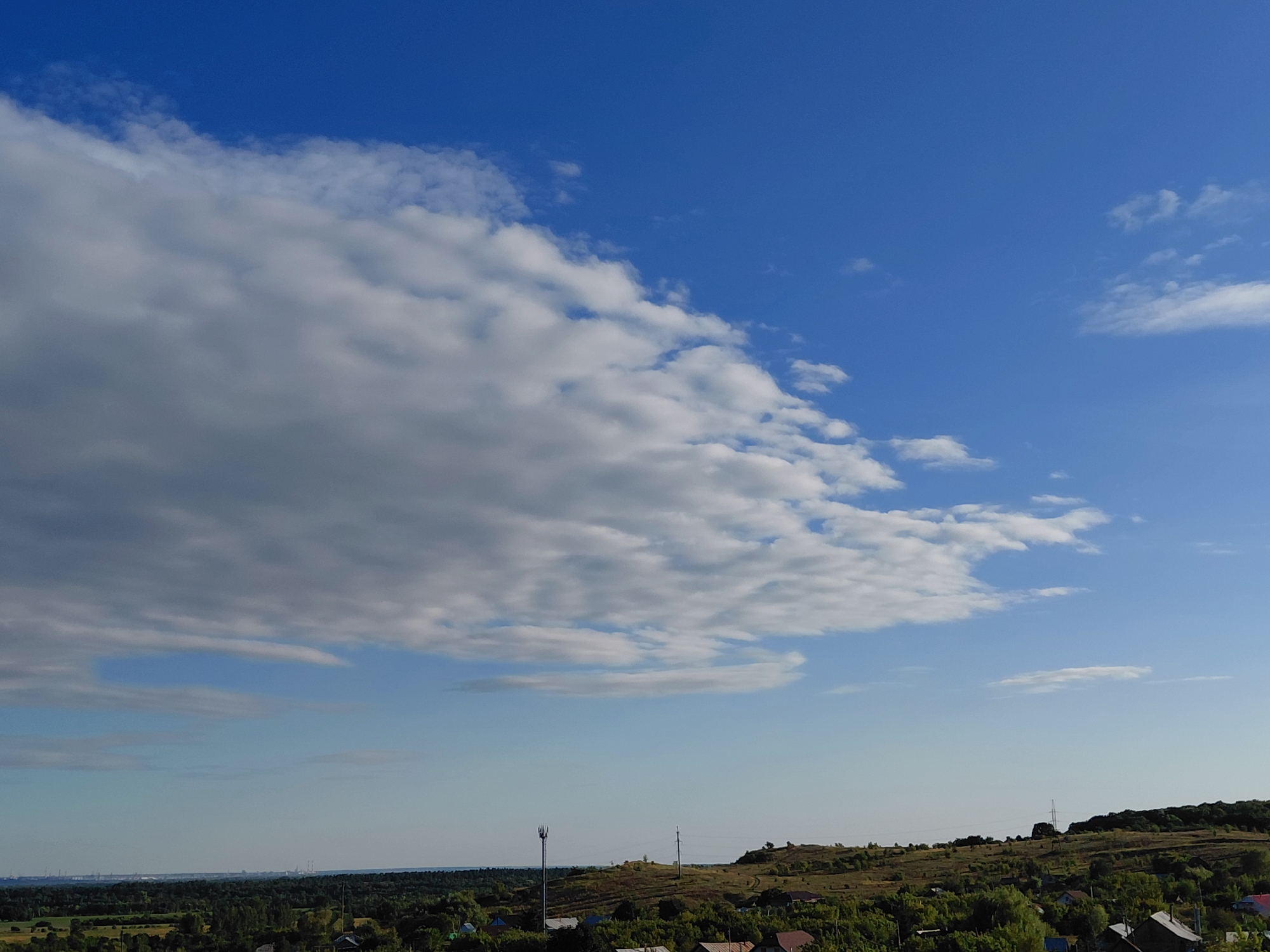 A wide-angle, eye-level, outdoor shot captures a vast expanse of the sky over the village of Verkhnyaya Podstepnovka, Russia. The sky is a vibrant blue, with large, fluffy white clouds dominating the upper left quadrant of the image and smaller scattered clouds across the rest of the sky. Below the sky, rolling hills covered in green trees and vegetation extend to the horizon. A few small houses with red roofs are visible nestled amongst the trees, indicating a rural or semi-rural setting. A tall cellular tower stands prominently on the left side of the frame, and a few more distant structures, possibly industrial, are faintly visible on the far left horizon. The overall scene is serene and peaceful, bathed in natural daylight under a clear sky.