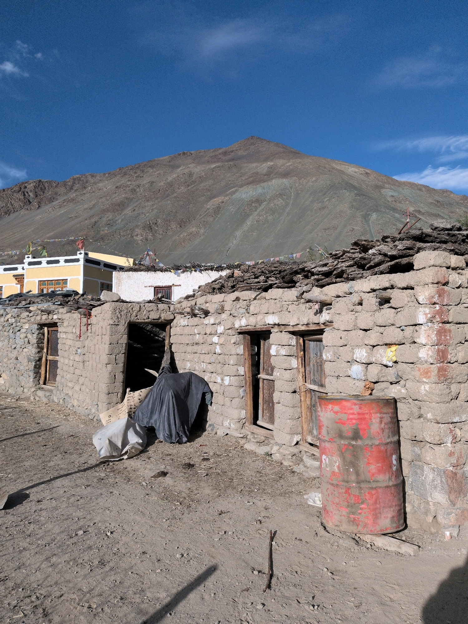 A low-angle, full shot captures a scene in Skidmang, India. In the foreground, a weathered, red barrel stands next to a mud-brick building with a roof made of layered stones and debris. The building has two open doorways and a wooden-framed window. Behind this structure, two more buildings are visible: one white and one yellow, both with Tibetan prayer flags draped across their roofs. The yellow building features a decorative white band along its top. In the background, a vast, barren mountain under a clear blue sky dominates the landscape, with a few wispy clouds scattered across the expanse. The ground is composed of dry dirt and small stones, with sparse vegetation.