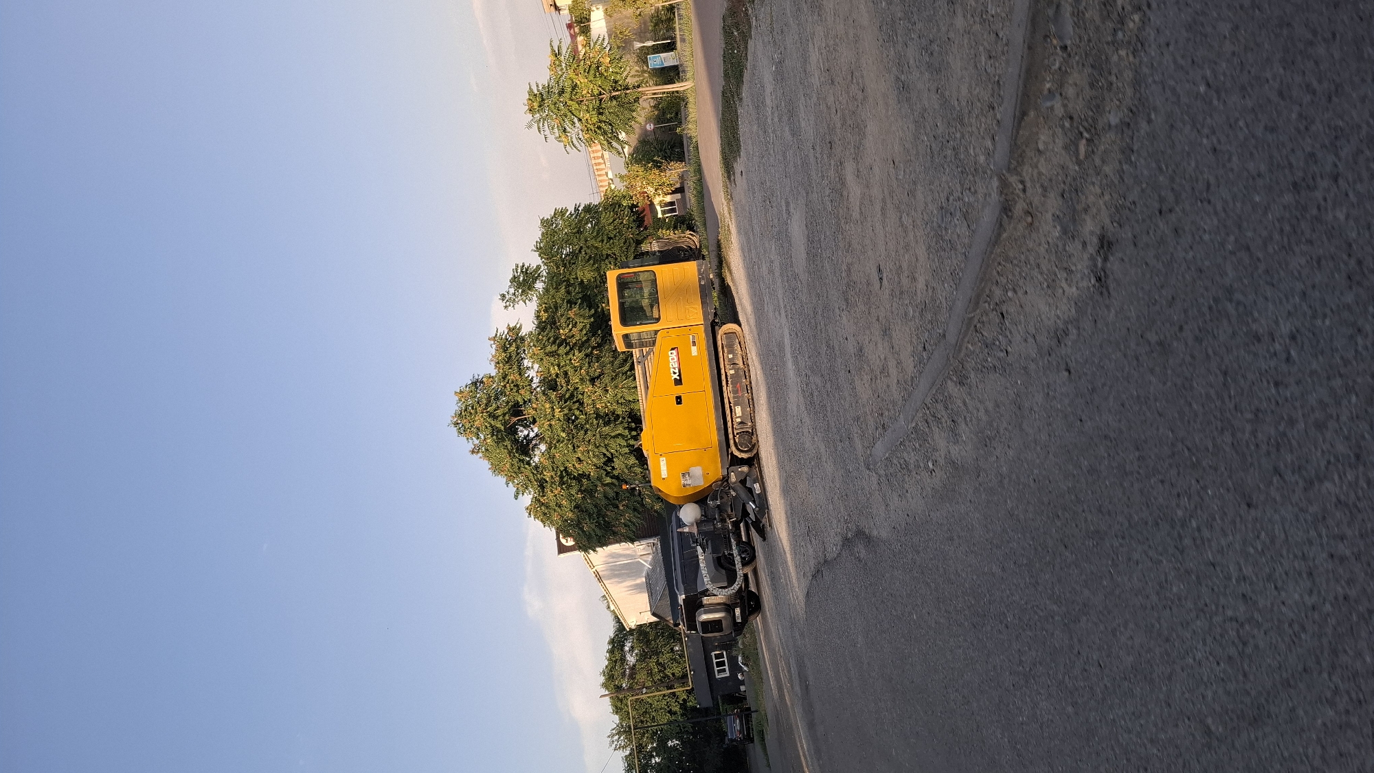 A bright yellow horizontal boring machine, marked with "XZ200," is parked on a dirt and gravel lot. The machine has tracks and a prominent operator's cab. It is positioned alongside a dark-colored SUV. Behind the vehicles, several large trees with lush green foliage are visible, and a white building with a dark roof is partially obscured by the trees. The sky above is clear blue with a few wispy clouds. The scene appears to be in a semi-urban or suburban area, potentially a construction or industrial site. The lighting suggests it is late afternoon or early evening, with the sun casting long shadows. The ground is a mix of paved and unpaved surfaces, indicative of an outdoor environment.