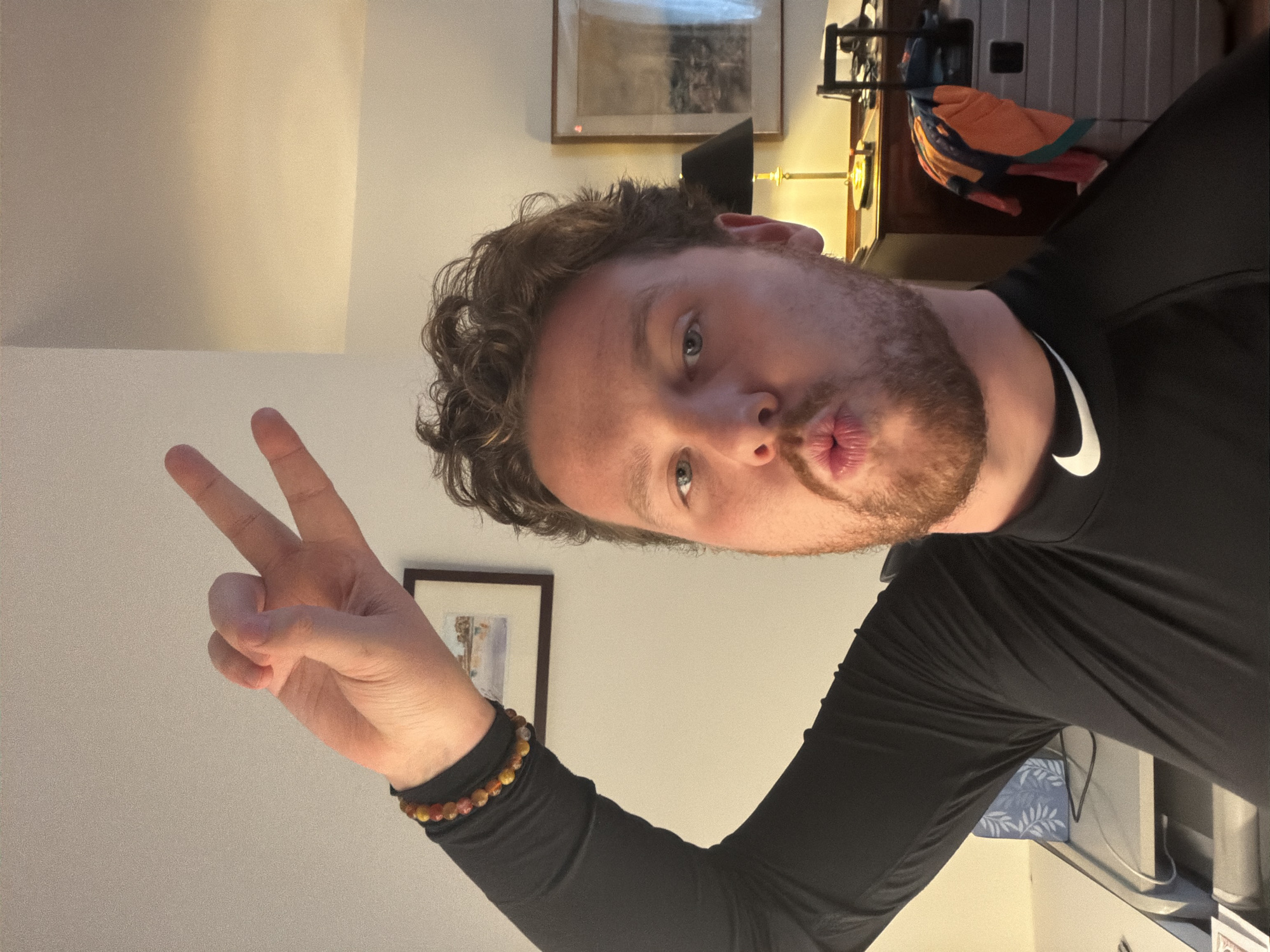 A man poses for a selfie indoors with a neutral white background. He has light skin, curly brown hair, and a short beard. He is wearing a long-sleeved black shirt with a white Nike swoosh on the neckline. The man is making a kissing face while holding up his left hand in a peace sign. He is also wearing a beaded bracelet on his left wrist.
Behind him on the wall are two framed pictures and a desk with a lamp. The location is presumably in a residential or office building, but not identifiable as any specific place in New York City.