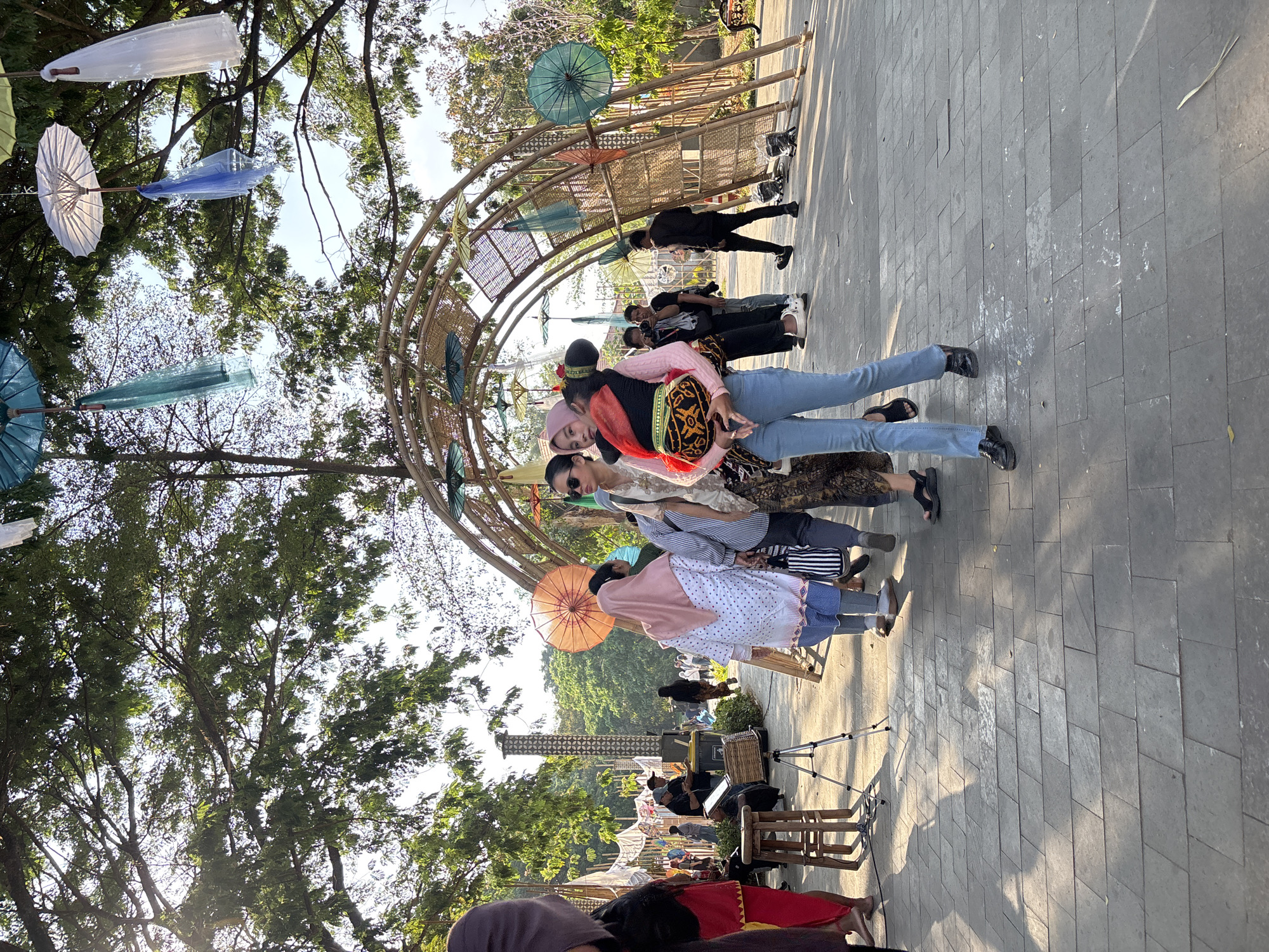 This image captures a lively scene at the Sriwedari Park in Surakarta, Indonesia, likely during an event. The foreground is dominated by a paved walkway with large, square gray tiles, casting long shadows from the overhead trees and structures.
In the midground, a group of people are gathered, some walking and others observing. A woman in the center, wearing blue jeans and a pink top, is carrying a child on her back. The child is dressed in a traditional outfit with an orange and black patterned fabric. Behind her, another woman in a pink headscarf and a white patterned top is walking away from the camera. To the left, a person in a dark outfit with red accents is partially visible.
The background features a large, arched bamboo structure decorated with numerous colorful umbrellas of various sizes and styles, some of which are translucent and appear to be made of fabric. Further back, more people are visible, as well as what appears to be a stage area with musical equipment and a tall, cylindrical structure with intricate patterns. Trees with lush green foliage provide a canopy over the scene, and the sunlight filtering through creates a warm, dappled effect. Several other decorative elements, like hanging fabrics and more umbrellas, are suspended from the trees. The overall atmosphere suggests a festive outdoor gathering or market.