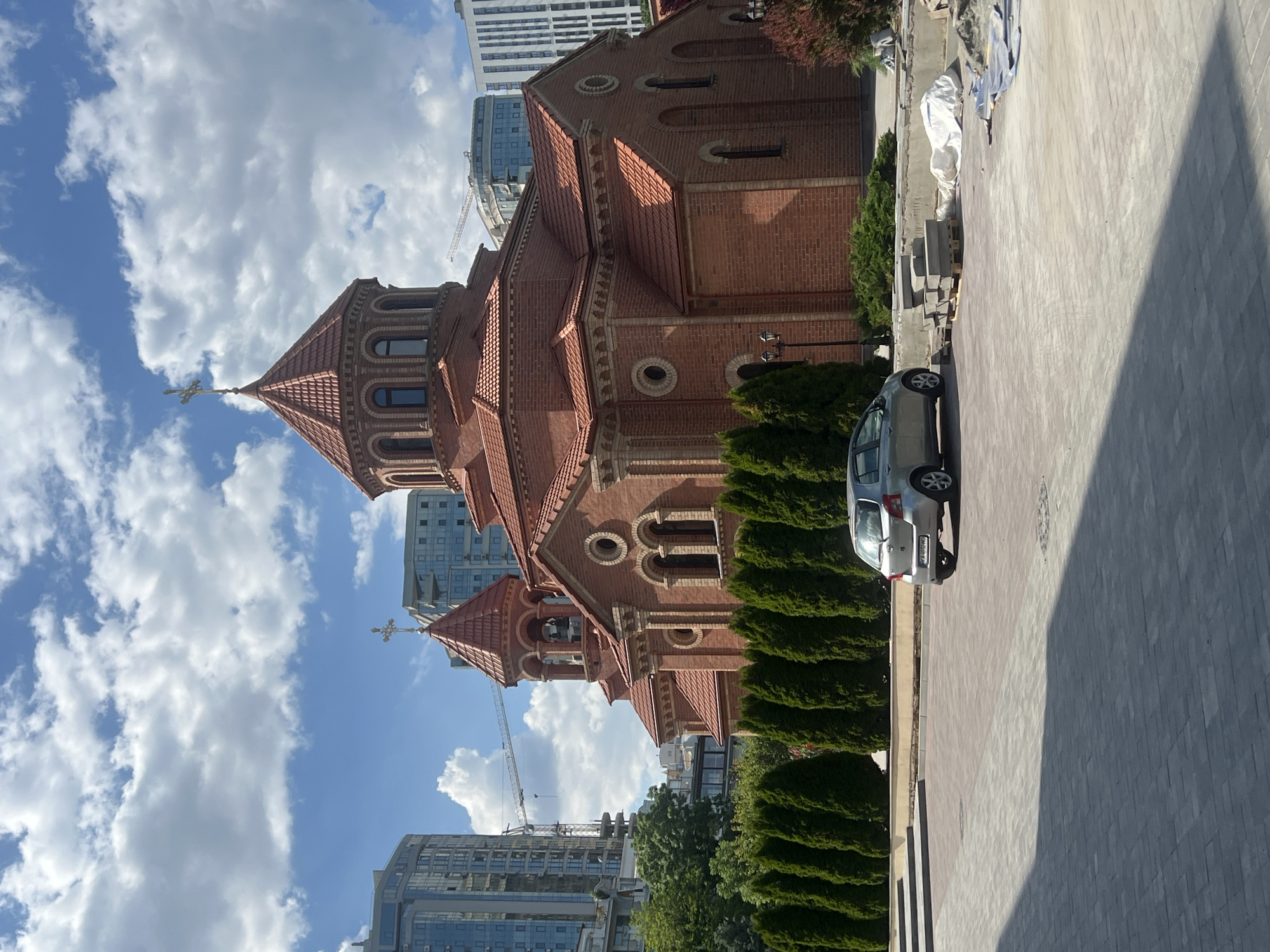 This is a medium-shot, eye-level photograph taken on a sunny day in Odesa, Ukraine. The primary subject is an Armenian Apostolic church constructed of red brick. The church features a prominent red-tiled dome topped with a cross and a smaller bell tower, also with a cross. In front of the church is a row of manicured green evergreen trees. To the left of the church, high-rise buildings are visible, suggesting an urban setting, and a construction crane is present. A silver hatchback car is parked in the foreground on a paved area. The ground is covered with interlocking pavers, and some construction materials, including pallets with stone slabs and white sheeting, are present on the right side of the image. The sky is blue with scattered white clouds.