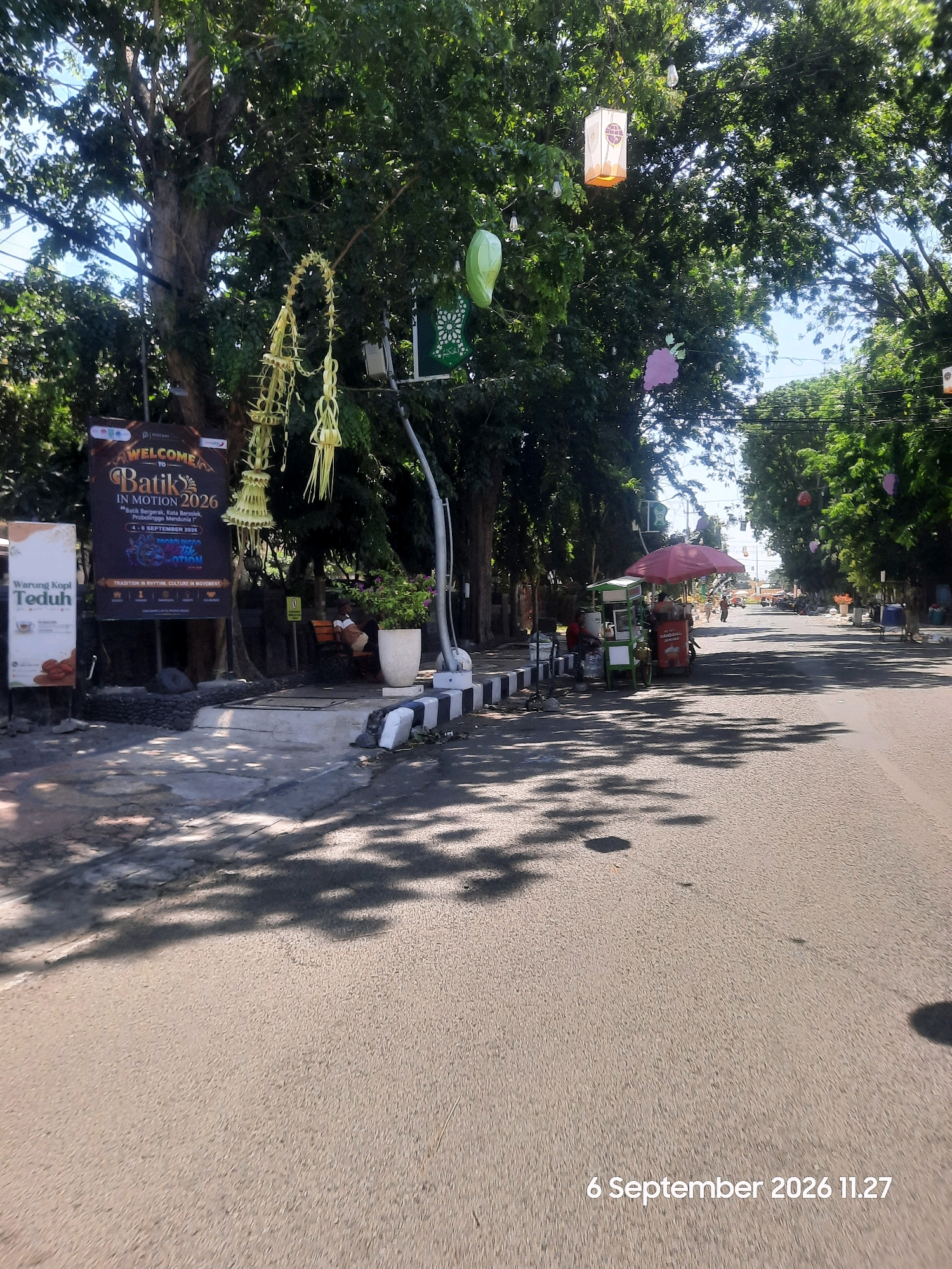 This is a medium shot of a street scene in Probolinggo, Indonesia, on September 6, 2026, at 11:27 AM. The image captures a section of a road lined with trees, with a sidewalk and some commercial establishments on the left.
On the left side of the road, there's a dark sign with text that appears to be an event advertisement. The sign has "WELCOME TO Batik IN MOTION 2026" prominently displayed. Below this, there's smaller text, potentially indicating event details or sponsors. Adjacent to this sign is another smaller white sign that reads "Warung Kopi Teduh".
Several decorative elements are hanging from the trees, including what look like woven palm leaf ornaments and colorful lanterns. One lantern is white with an orange glow, and another is green and bulbous. Further down the street, a pink umbrella shades a food cart, and a person is seen behind it. Another cart, possibly a food or drink vendor, is parked next to it.
Further down the road, there are other street vendors and parked vehicles, indicating a bustling commercial area. The road surface is asphalt, and dappled sunlight casts shadows from the trees onto the pavement. A few people are visible in the distance, walking along the street or sitting on benches. The overall environment suggests a warm, sunny day in an urban setting.