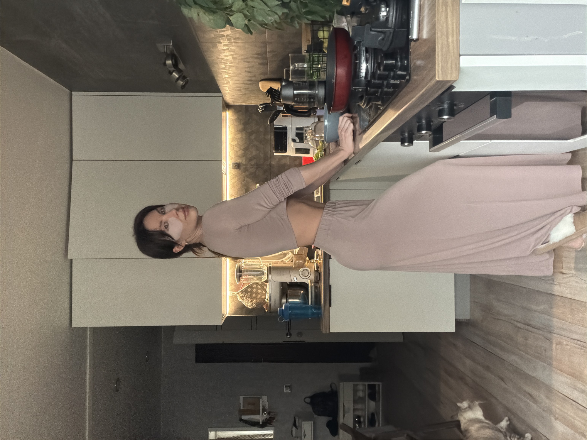 A woman with dark hair is standing in a kitchen in what appears to be an apartment in Perm, Russia. She has a light-colored patch on her face under her left eye and is wearing a light brown two-piece outfit that exposes her midriff. She is reaching towards kitchen appliances or items on the countertop.
The kitchen features light gray cabinets, a dark gray ceiling, and wood-patterned flooring. There are various kitchen appliances, including a blender and a cooktop. In the background, there's indirect lighting and a view into another part of the apartment. A cat is lying on the floor.
