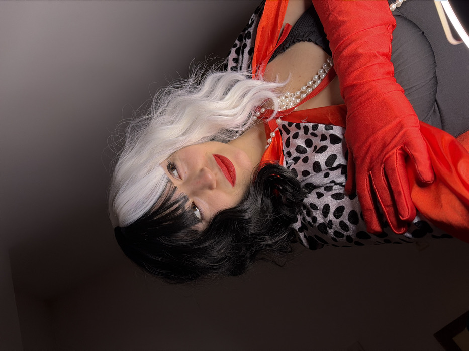 A female subject, styled to resemble Cruella de Vil, is depicted from a low-angle perspective. She wears a prominent half-black, half-white wig with bangs, red lipstick, and eyeliner. Her gaze is directed upwards and to the right, displaying a subtle smile. She is attired in a faux dalmatian-print coat or cape, layered over a red collar or trim, and a multi-strand white pearl necklace. Long red gloves cover her forearms, with her gloved hands positioned prominently in the lower-right foreground. A black garment is visible beneath her outer attire. The background is uniformly dark and indistinct, indicating an interior setting with localized lighting. The precise location within Budva, Montenegro, is not discernible from the visual evidence.