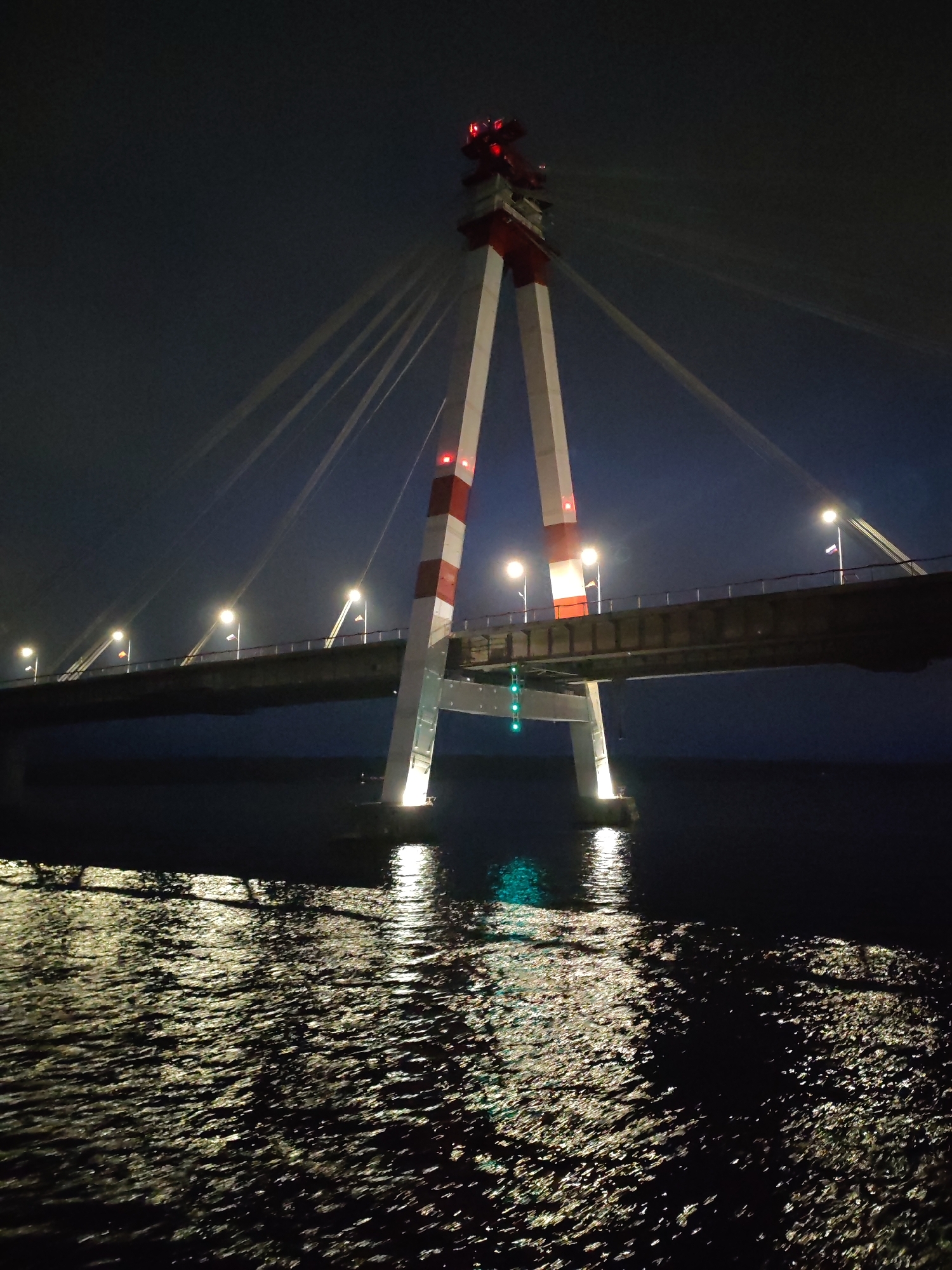 At night, a cable-stayed bridge, likely spanning a body of water, is prominently featured. The bridge's central support towers are striped in red and white, with red lights at the top. Cables extend from the towers to the bridge deck. Streetlights line the bridge deck, and several flags are visible. The water surface shows reflections of lights and the bridge structure. A series of green lights are seen beneath the bridge deck on one of the support structures. The overall setting is dark, emphasizing the illuminated structure.