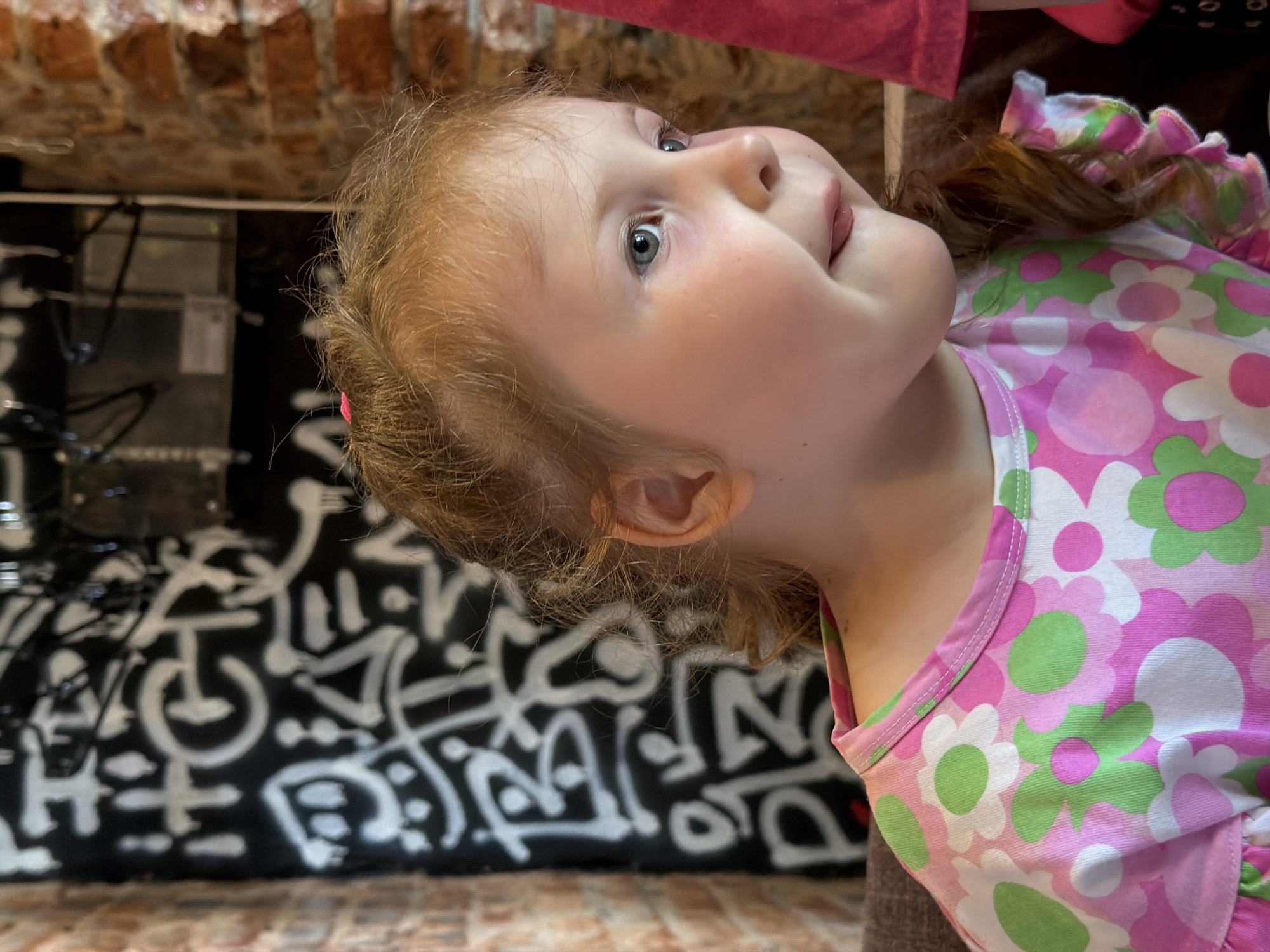This image, captured in the city of Chaikovsky, Russia, features a young child in a close-up, right-profile view. The child has fair skin, rosy cheeks, and light blue-grey eyes gazing towards the right, off-camera. Her reddish-blonde hair, styled with some waves or curls, is partially gathered, with a small pink hair tie visible near the crown of her head. Her lips are slightly parted, revealing the tip of her tongue, suggesting a curious or slightly amused expression. She is wearing a light-colored top or dress patterned with large pink, green, and white flowers, featuring ruffled sleeves. The background consists of a dark wall on the left adorned with extensive white, stylized graffiti or tagging that is illegible. To the left and right sides, sections of exposed reddish-brown brickwork are visible. In the upper background, dark, indistinct structural elements are present. A portion of another person, clad in a pink garment, is partially visible on the far right.
