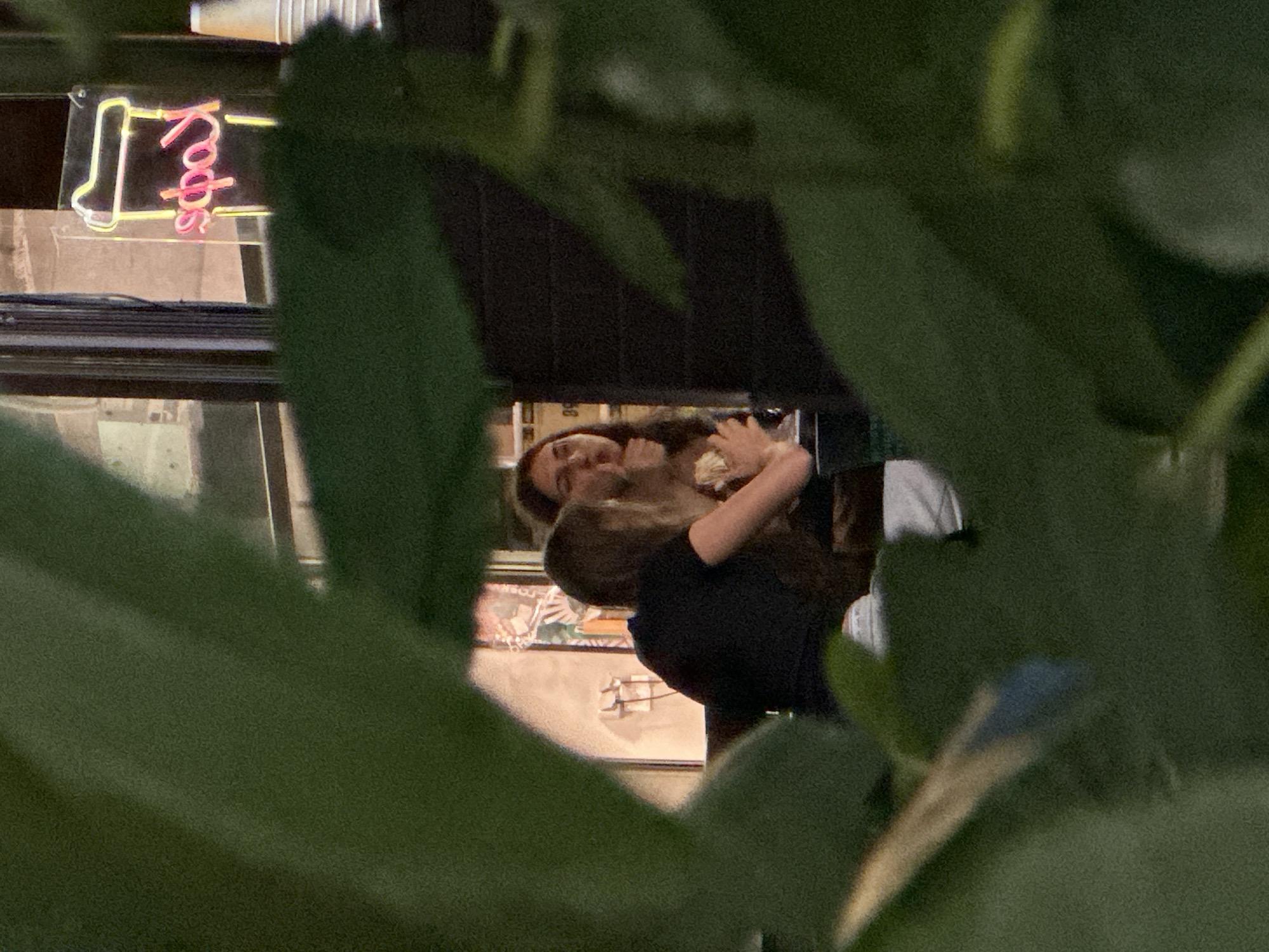 This is a slightly blurry, medium-shot, outdoor photograph taken at night, possibly through foliage, of two young women sitting at a table. The woman on the left is wearing a black t-shirt and is looking towards the right. The woman on the right is wearing a light-colored top and is looking down and to her left, with her hands near her face, possibly eating. In the background, through a window, a neon sign shaped like a coffee cup with the Russian word "Кафе" (Cafe) written on it in pink and yellow is visible. There are also some stacked white paper cups on a shelf to the right of the neon sign. The setting appears to be a cafe or restaurant in the city of Sochi, Russia. The overall impression is that the photograph was taken discreetly, with the foliage obscuring some of the view.