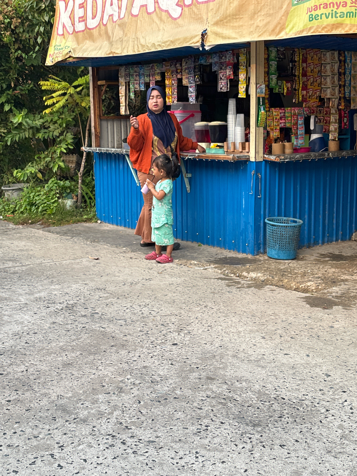 A woman wearing a dark blue hijab and an orange cardigan stands at a blue corrugated metal kiosk in Banjarmasin, Indonesia. She is looking to her left and holding a small pink cup in her right hand. Beside her, a young girl in a mint green dress with a floral pattern stands holding a similar pink cup. The kiosk is stocked with various packaged goods, and there is a blue plastic trash can to the right of the kiosk. The ground is paved with asphalt, and there is greenery and trees in the background. The kiosk has a yellow awning with text that appears to be in Indonesian.