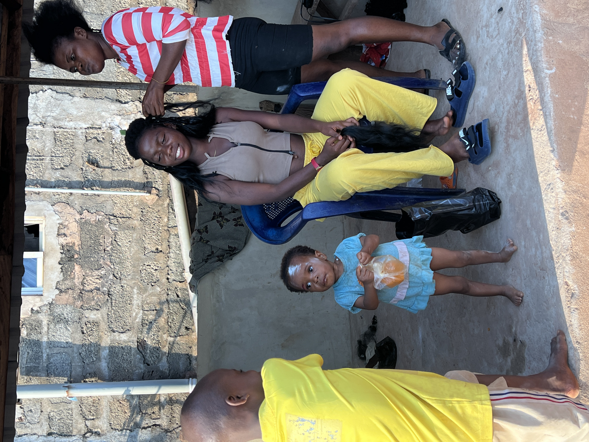 The media file shows an outdoor scene in Nkpor, Nigeria, with several people and objects. In the foreground, a person wearing a bright yellow shirt is lying down, with their legs extended. To their right, a young child in a blue dress is sitting and holding a plastic bag containing orange objects. Behind them, two young women are engaged in braiding hair. One woman, wearing a red and white striped shirt and black shorts, stands and is actively braiding the hair of another woman who is seated. The seated woman is wearing a light-colored tank top with a zipper and bright yellow pants. The background reveals a textured, weathered wall and the ground appears to be concrete or packed earth. The lighting suggests it is daytime, likely sunny. The overall atmosphere appears casual and domestic, possibly a family or friends gathering.