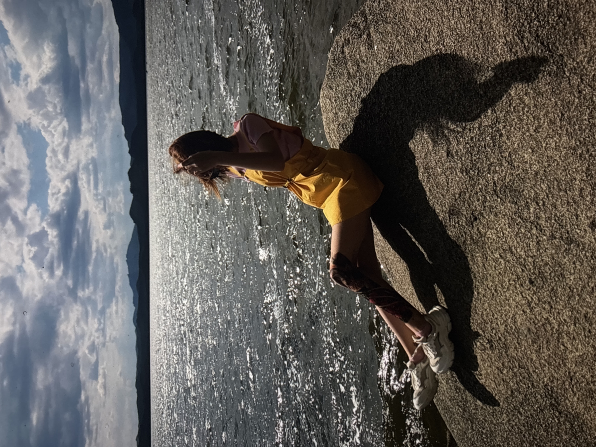 An individual with reddish-brown hair sits on a large rock by a body of water, with mountains in the background under a cloudy sky. The individual is wearing a pink shirt, a yellow dress, and white sneakers. The individual's left leg appears to have a dark, patterned covering or tattoo extending from the knee down. The sun casts a shadow of the individual onto the rock. The body of water is choppy and reflects the sunlight. There are no discernible Omsk landmarks or features present in the image.