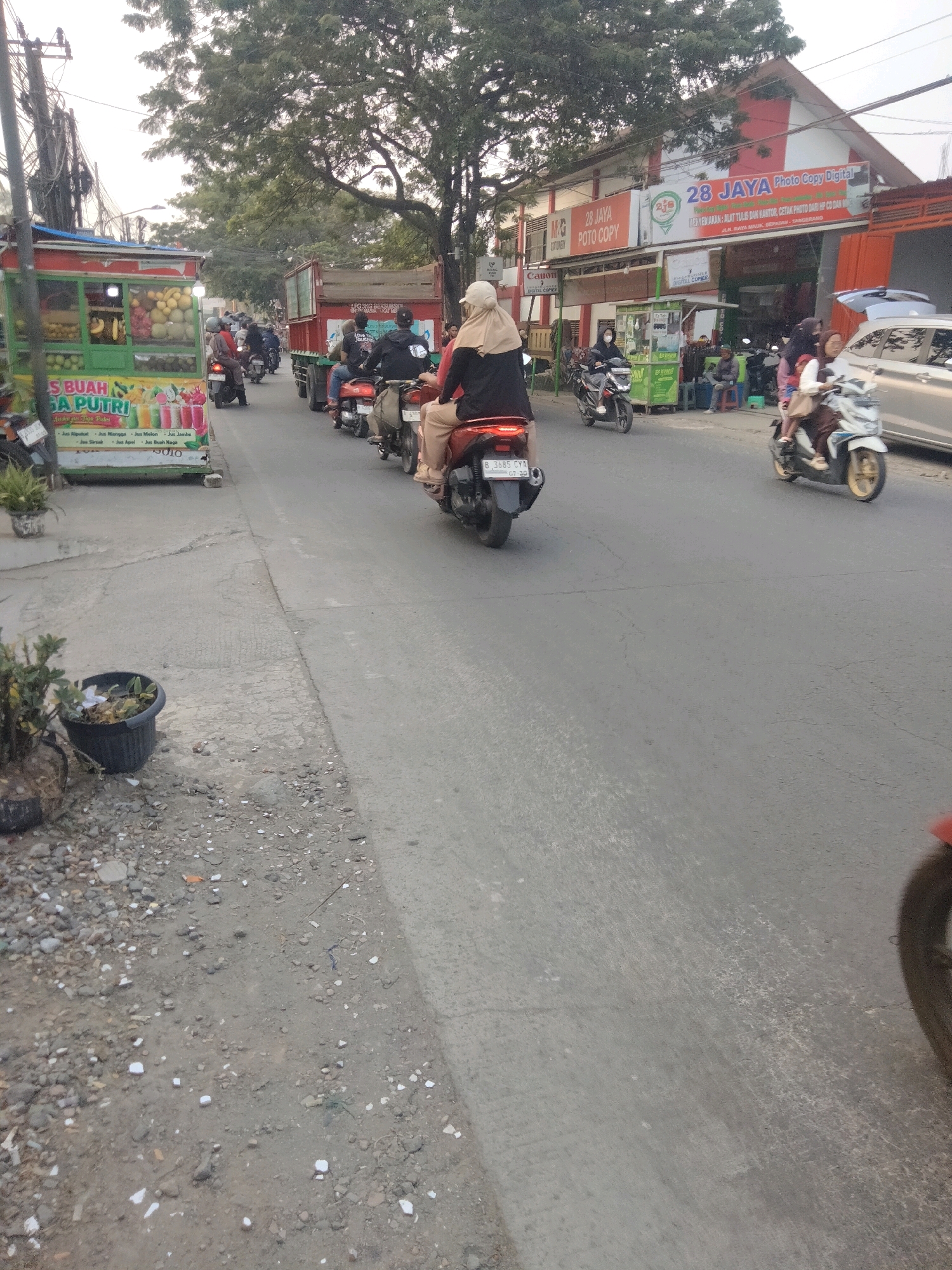 In Sepatan, Indonesia, on a paved road lined with businesses, a fruit juice cart vendor is parked on the left side of the street, displaying various fruits and colorful juice advertisements. A red truck is visible behind the cart, heading away from the viewer. Several motorcycles are proceeding down the road in the same direction, with riders wearing helmets and casual attire. One motorcycle in the foreground on the right is white with a yellow rim, carrying a rider. A second motorcycle on the left, closer to the cart, is dark colored and also carries a rider. The environment appears to be a commercial area with buildings in the background, including a sign for "28 JAYA Photo Copy Digital." The sky is overcast, and the overall lighting suggests it is daytime.
