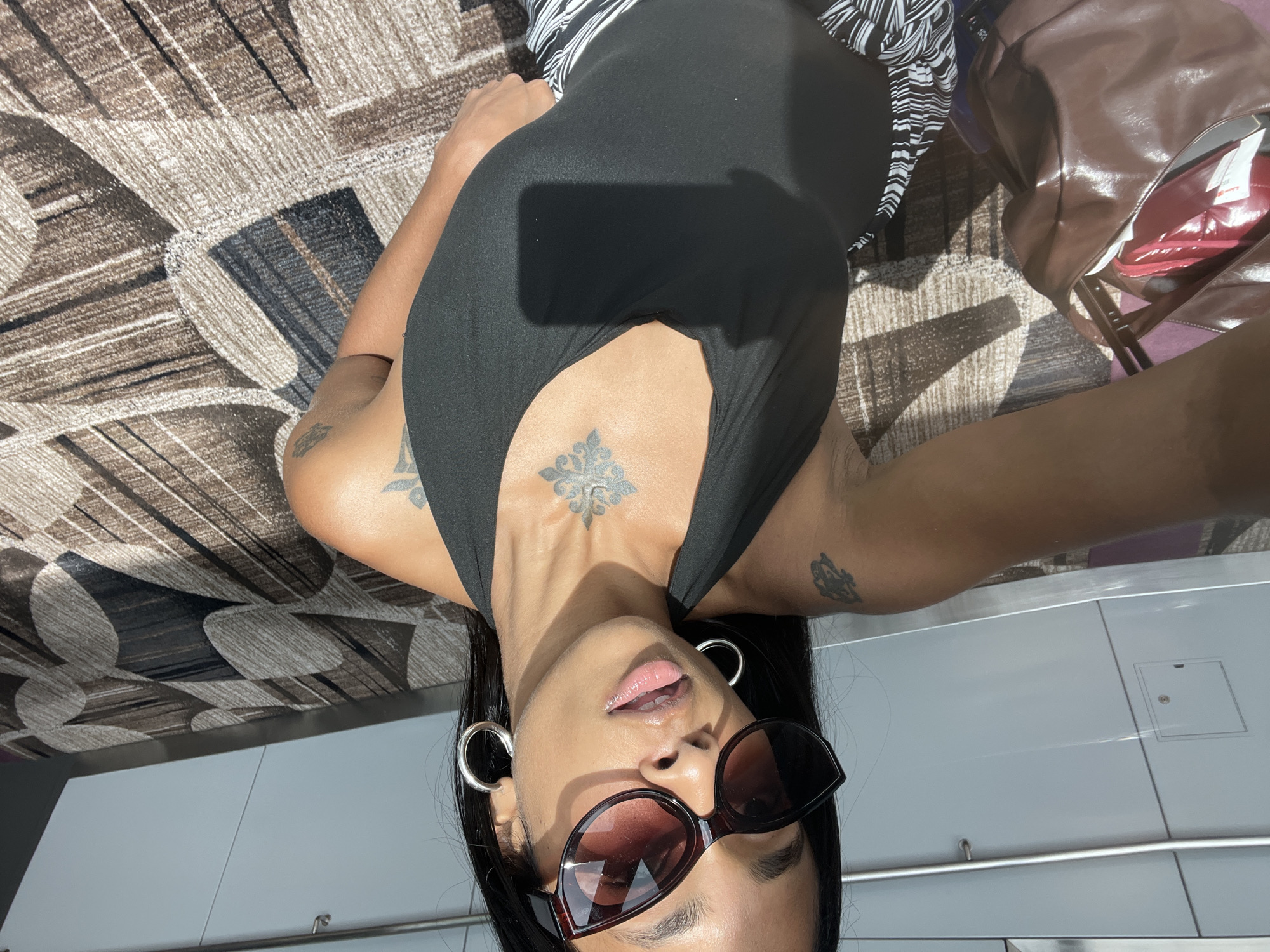 A woman with dark hair and sunglasses is taking a selfie in Singapore, Singapore. She is wearing a black halter top and a striped skirt. She has several tattoos on her arms and chest. In the background, there is a patterned carpet and a wall with white panels and a metal railing. A brown handbag is visible on the left side of the image. The lighting suggests it is daytime, with sunlight casting shadows on the carpet.