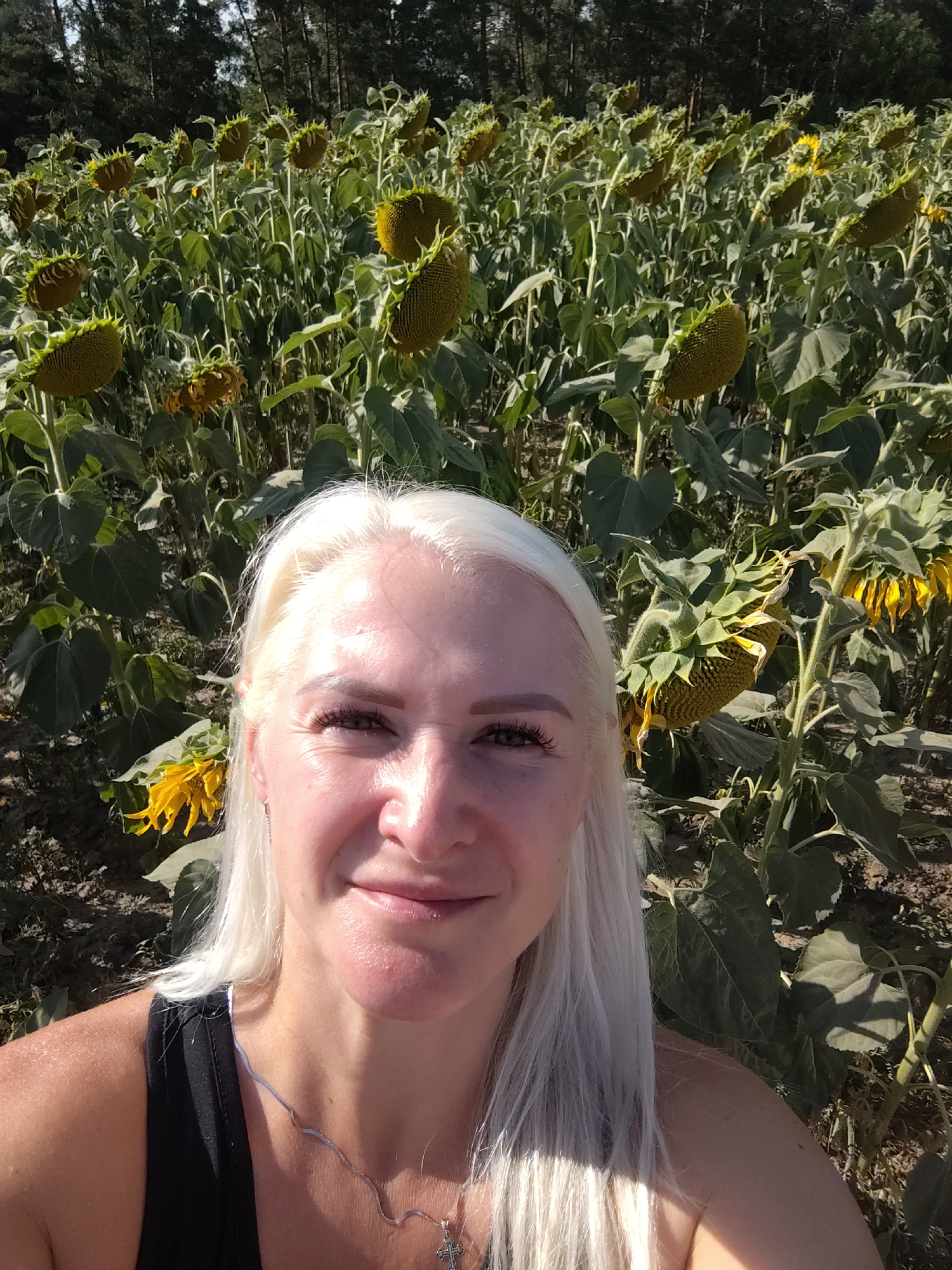 A blonde woman is taking a selfie in a sunflower field. She is smiling and looking at the camera. The field is full of sunflowers, and the trees are in the background. The image was taken in Balyko-Shchuchynka, Ukraine.