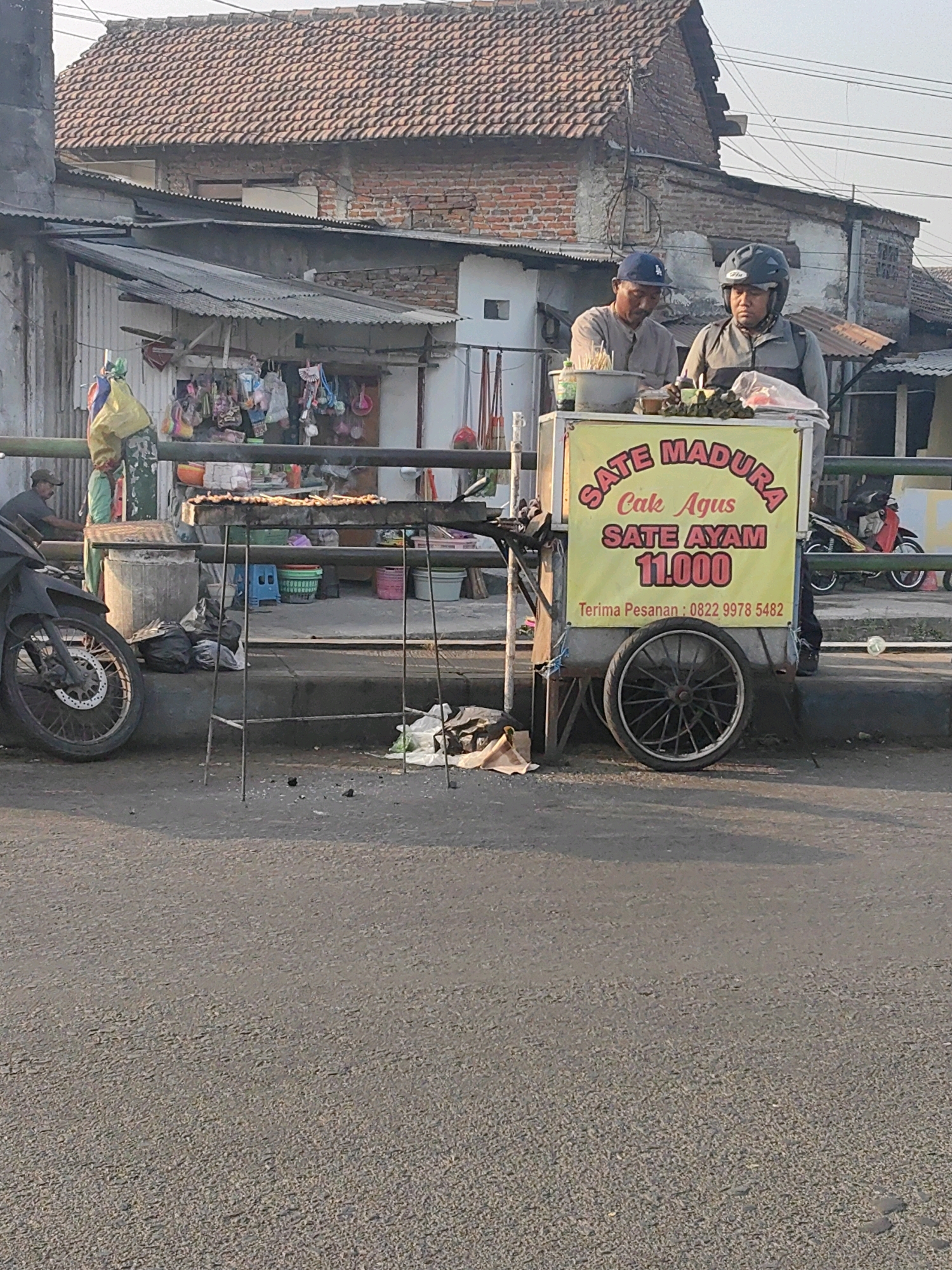The image shows a street scene in Keboansikep, Indonesia, with a food cart prominently displayed. Two men, one wearing a blue baseball cap and the other a black helmet, stand behind the cart. The cart has a yellow banner with red lettering that reads "SATE MADURA Cak Agus SATE AYAM 11.000 Terima Pesanan: 0822 9978 5482". Grilled skewers of meat are visible on a metal rack above the cart. In the background, there are buildings with tiled roofs and corrugated metal awnings, and small shops selling various items. A black motorcycle is parked to the left of the cart, and several black garbage bags are piled on the ground. The scene appears to be set during the late afternoon, with sunlight casting shadows on the road.
