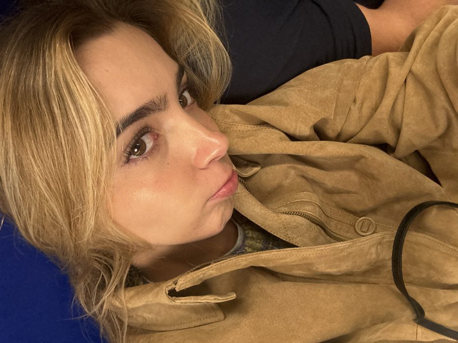 A female individual with blonde hair and fair skin is depicted in a close-up, head-and-shoulders portrait. Her head is tilted to the left, resting against an unseen dark blue or black surface, with her gaze directed forward towards the viewer. She has brown eyes, defined eyebrows, and appears to be wearing mascara and a light lip gloss on her slightly pouting lips. She is dressed in a tan or light brown suede-like jacket, featuring a collar, a visible zipper running down the front, and a circular snap button closure on the lower front left. A segment of a patterned garment, possibly a green, yellow, and grey sweater, is visible beneath the open jacket collar. The upper left background reveals a solid blue surface. A black strap is visible in the lower left foreground. The specific location within the city of Best, Netherlands, or any other location, cannot be determined from the visual information provided in the image.
