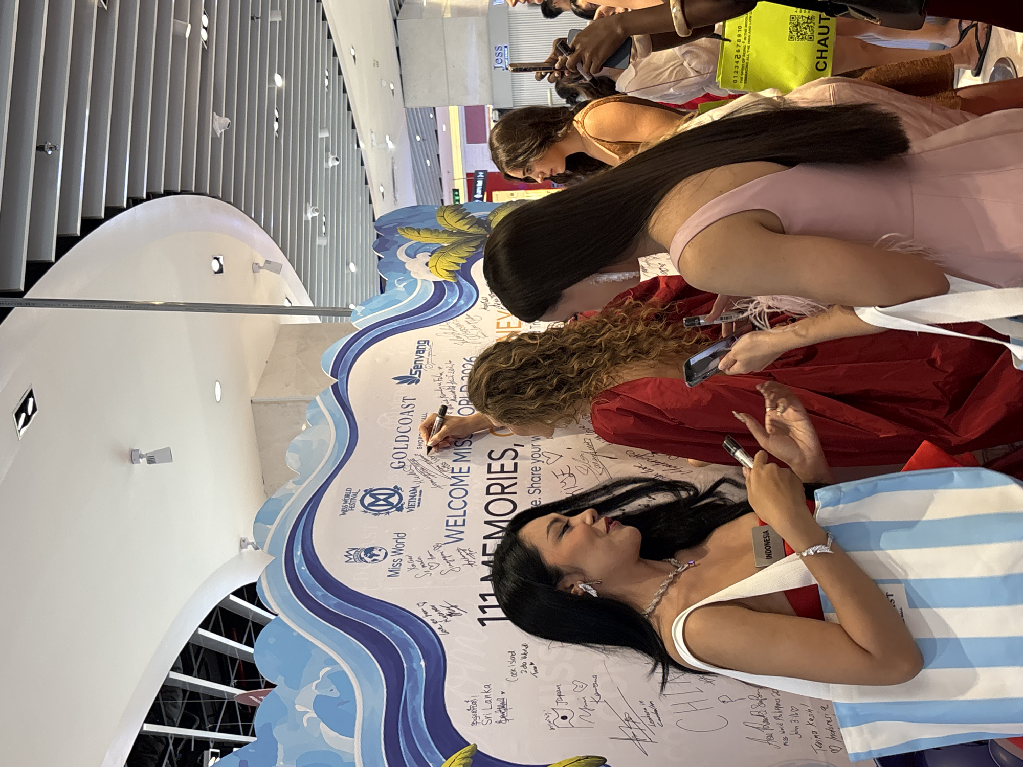 The image shows a section of a decorative white wall with blue wave motifs and palm tree graphics. The wall is covered with numerous handwritten signatures and messages, indicating it is a commemorative display or guest book. The words "WELCOME MISS" are prominently displayed in large letters, with "111 MEMORIES" below. Several people are visible interacting with the wall and each other.
In the foreground left, a woman with long dark hair, wearing a red top and a necklace, holds a marker and a blue and white striped tote bag. She is looking towards her right, interacting with another woman. She has a name tag that reads "INDONESIA".
In the center, a woman with long curly blonde hair, wearing a red dress, is actively writing on the wall with a marker.
To the right of the blonde woman, a woman with long dark hair, wearing a pale pink dress with feather accents, is looking at her phone. She is holding a white tote bag in her left hand.
Other individuals are partially visible in the background, suggesting a public event or gathering. The setting appears to be indoors, possibly a shopping mall or a convention center, with ceiling lights and structural elements visible above. There is no indication of the city of Nha Trang, Vietnam, in the visual information provided.