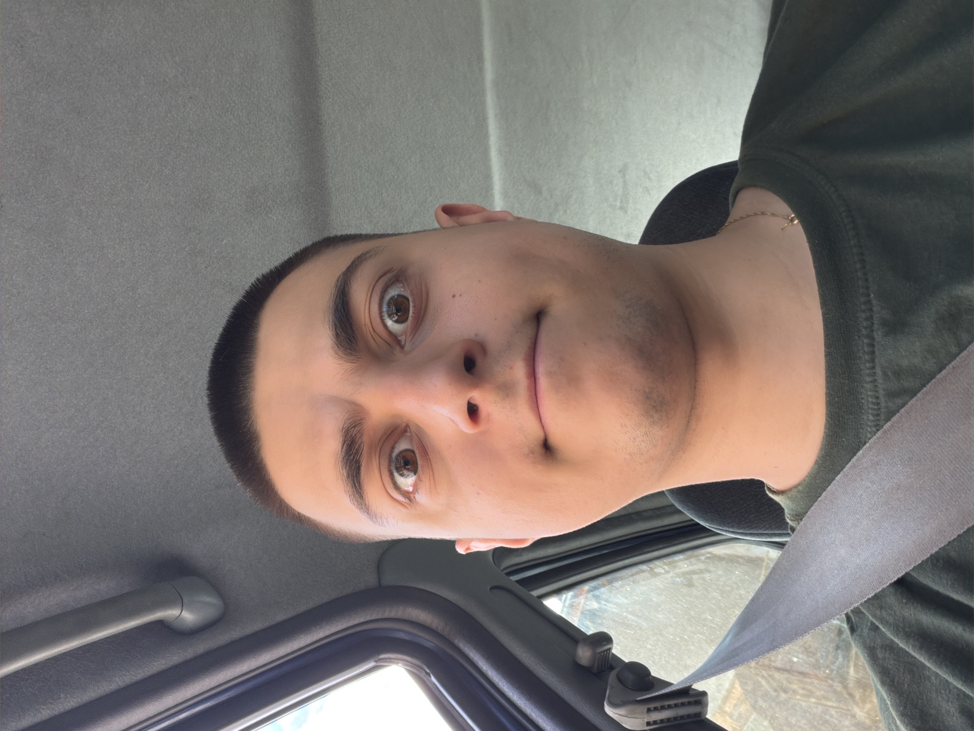 A close-up, selfie-style shot inside a car shows a person wearing a dark green shirt and a seatbelt. The car's interior includes a gray headliner, a grab handle, and a portion of the windshield. The person has short, dark hair, brown eyes, and light skin. A gold chain is visible around their neck. The exact location in the city of Tran, Bulgaria, cannot be determined from the image.