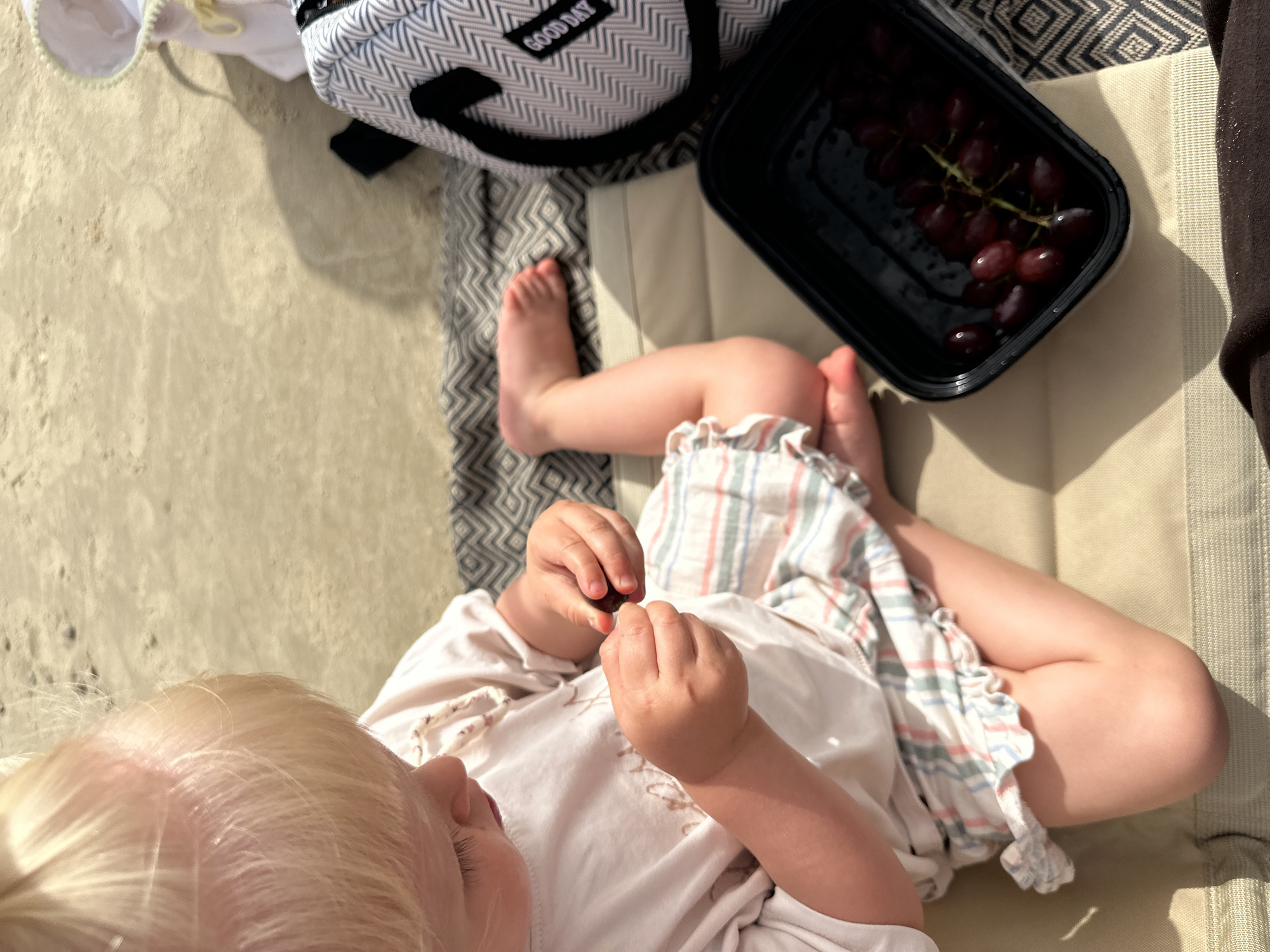 A fair-skinned toddler with blond hair sits on a beige beach mat in Tel-Aviv, Israel. The child wears a white shirt and striped shorts. The toddler’s bare feet extend from under the shorts. The child is holding a red grape in their hands. A black container of red grapes rests on the beach mat beside the child’s legs. A gray and white cooler with “GOOD DAY” printed on it sits to the left of the child.