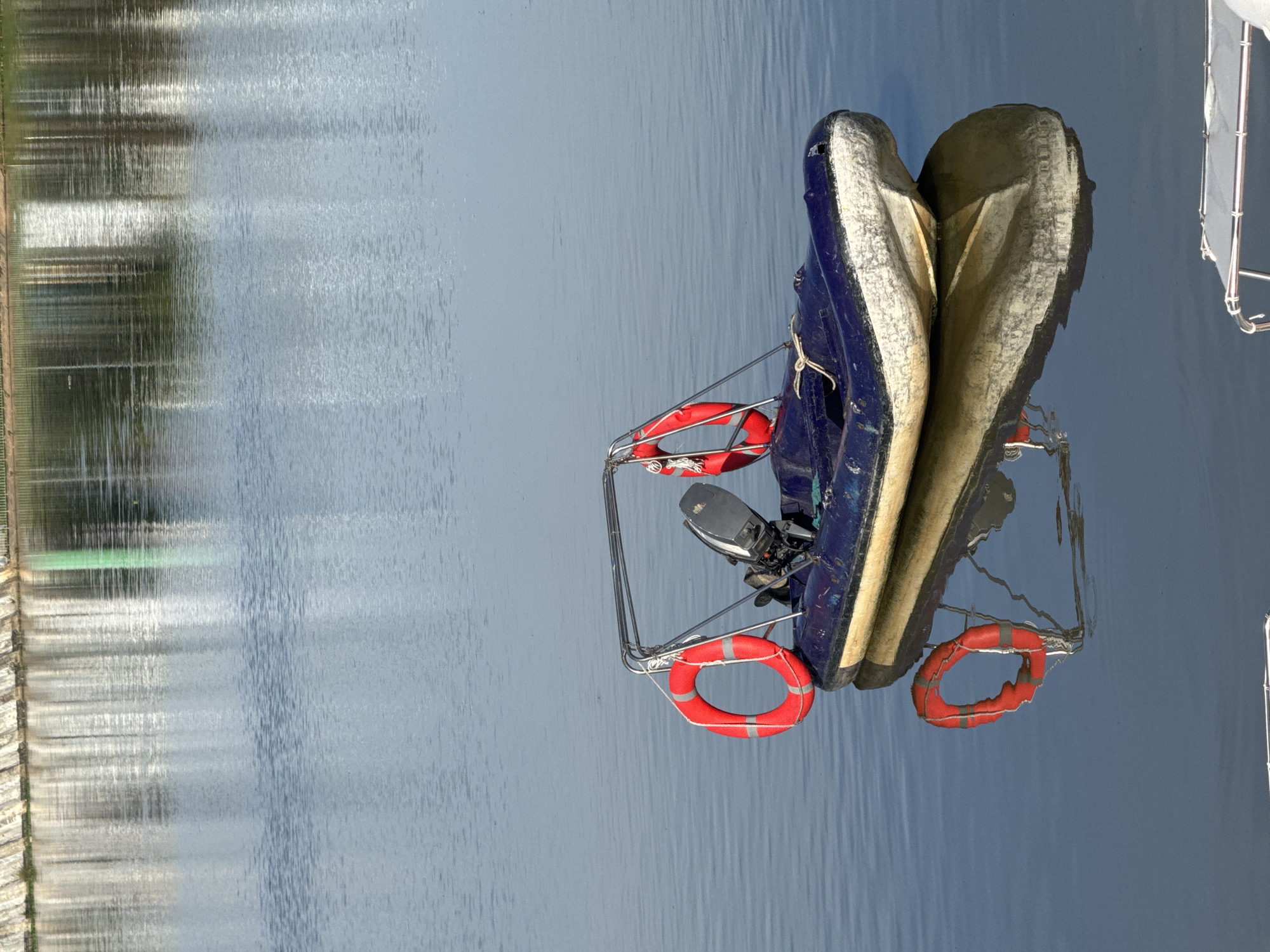The image shows an inflatable boat hanging from a davit, likely on the side of a larger boat, over calm water. The boat is blue and beige, with a small outboard motor attached. Several red lifebuoys are hanging from the davit. The water reflects the surrounding environment, including trees and buildings, suggesting a lake or harbor location. Based on the provided information, the location is Da Lat, Vietnam.