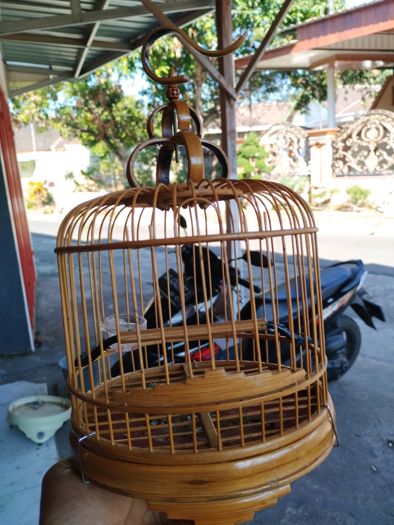 The image, taken in Sumberpetung, Indonesia, displays a close-up of a handmade bamboo birdcage held by a person's hand. The birdcage is intricately woven and has a natural bamboo finish, with a decorative hook at the top. Inside the cage, a small, clear plastic water dispenser with some green matter is visible. The cage is positioned in front of a partially obscured black motorcycle. The background shows a typical Indonesian street scene with a covered structure, greenery, and parts of a building. The lighting suggests it is daytime.