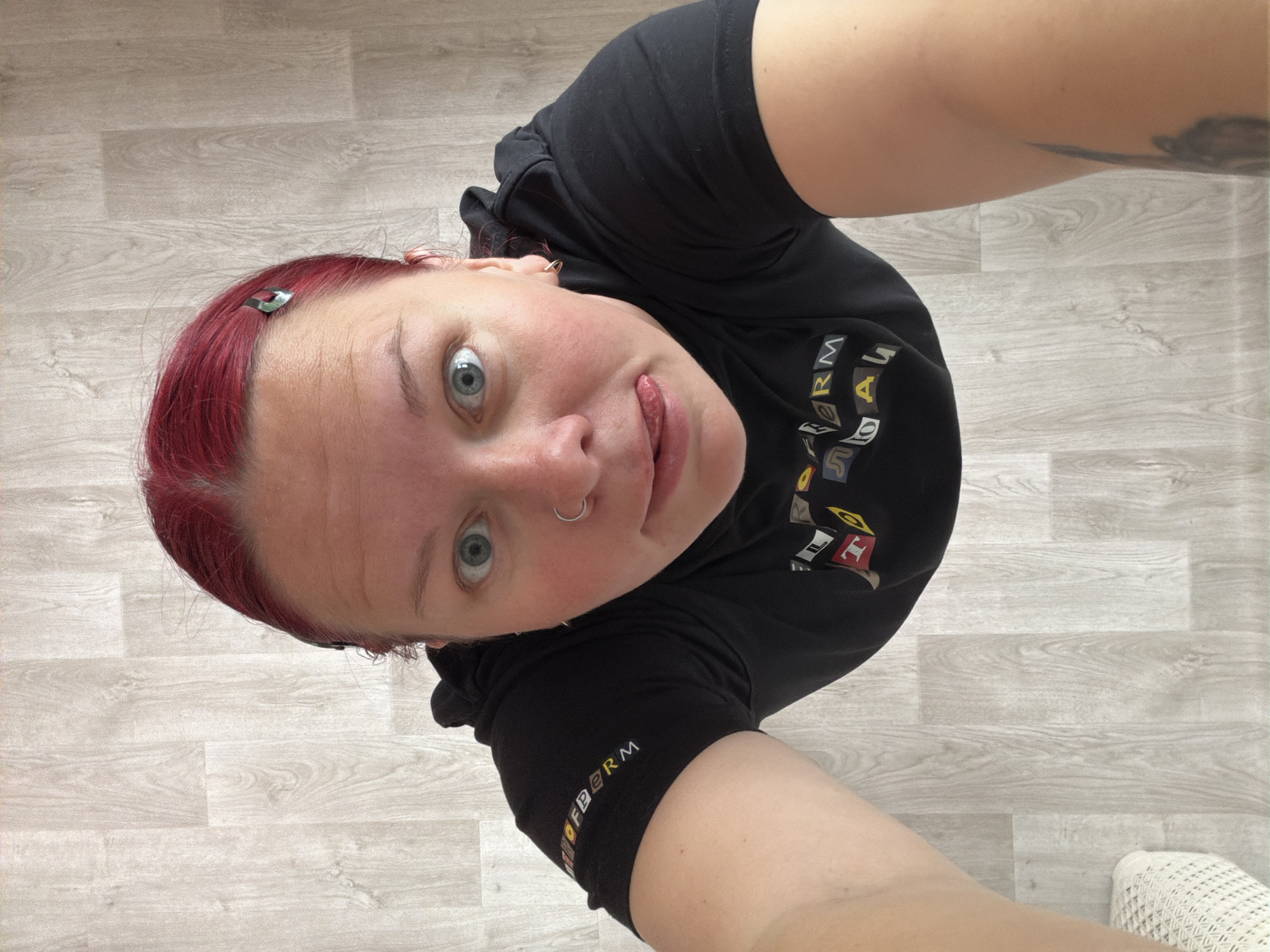 A selfie taken from above looking down at a woman with red hair and blue eyes. She has a nose ring and a silver hair clip. She is wearing a black t-shirt with lettering on the front. She is sticking her tongue out. The background is a light-colored wood-patterned floor. Part of a white shoe is visible in the lower right corner. There is a tattoo on her arm. No information is available about this image's location in the city of Chaikovsky, Russia.