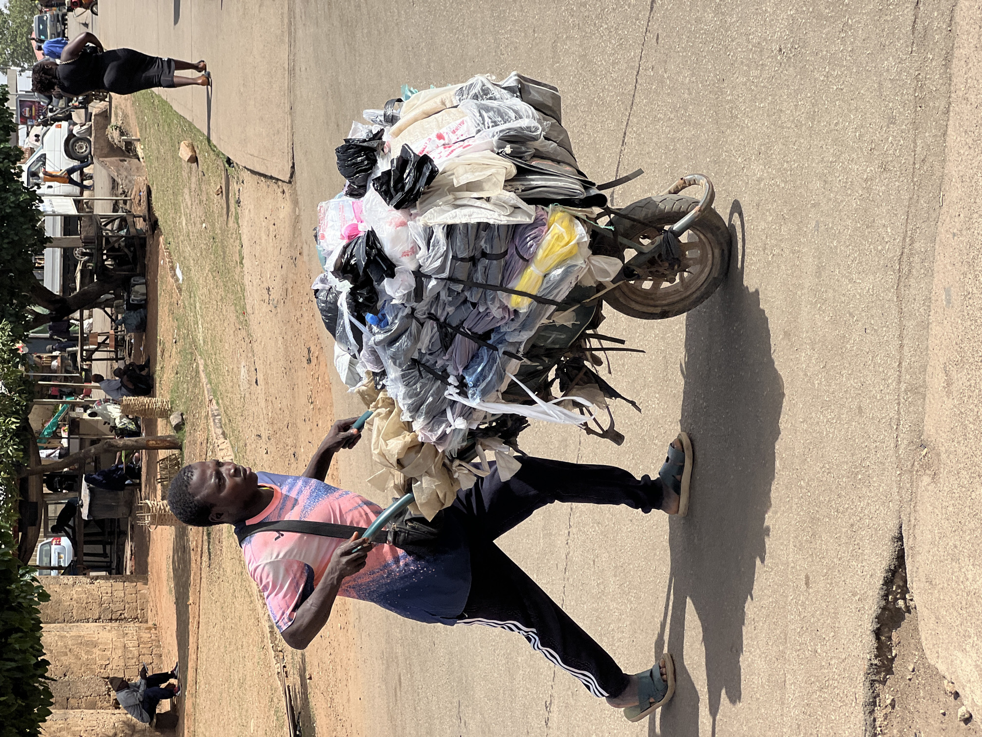 The photograph captures a street scene under bright, clear daylight, likely in an urban or semi-urban area. In the foreground, a man pushes a heavily laden two-wheeled cart on a paved road. He wears a short-sleeved t-shirt with a pink and blue abstract pattern, dark athletic pants with white stripes, and light-colored sandals. His dark hair is short. The cart is piled high with a large assortment of goods, primarily packaged in various colored plastic bags (black, white, clear, yellow, and purple) and secured with what appear to be straps. The load is significantly taller and wider than the cart itself.
To the left and behind the man, there is a dirt patch with sparse green vegetation, leading to a more bustling background. Here, several people are visible, some standing and some sitting, amidst what appears to be an informal market or residential area with simple structures and tree foliage. Further down the paved road, a woman in a black dress walks away from the viewer. Parked vehicles, including white vans, can be seen in the mid-ground. Strong shadows cast by the man and the cart suggest it is midday or early afternoon. No discernible text is clearly visible on any of the objects or subjects.