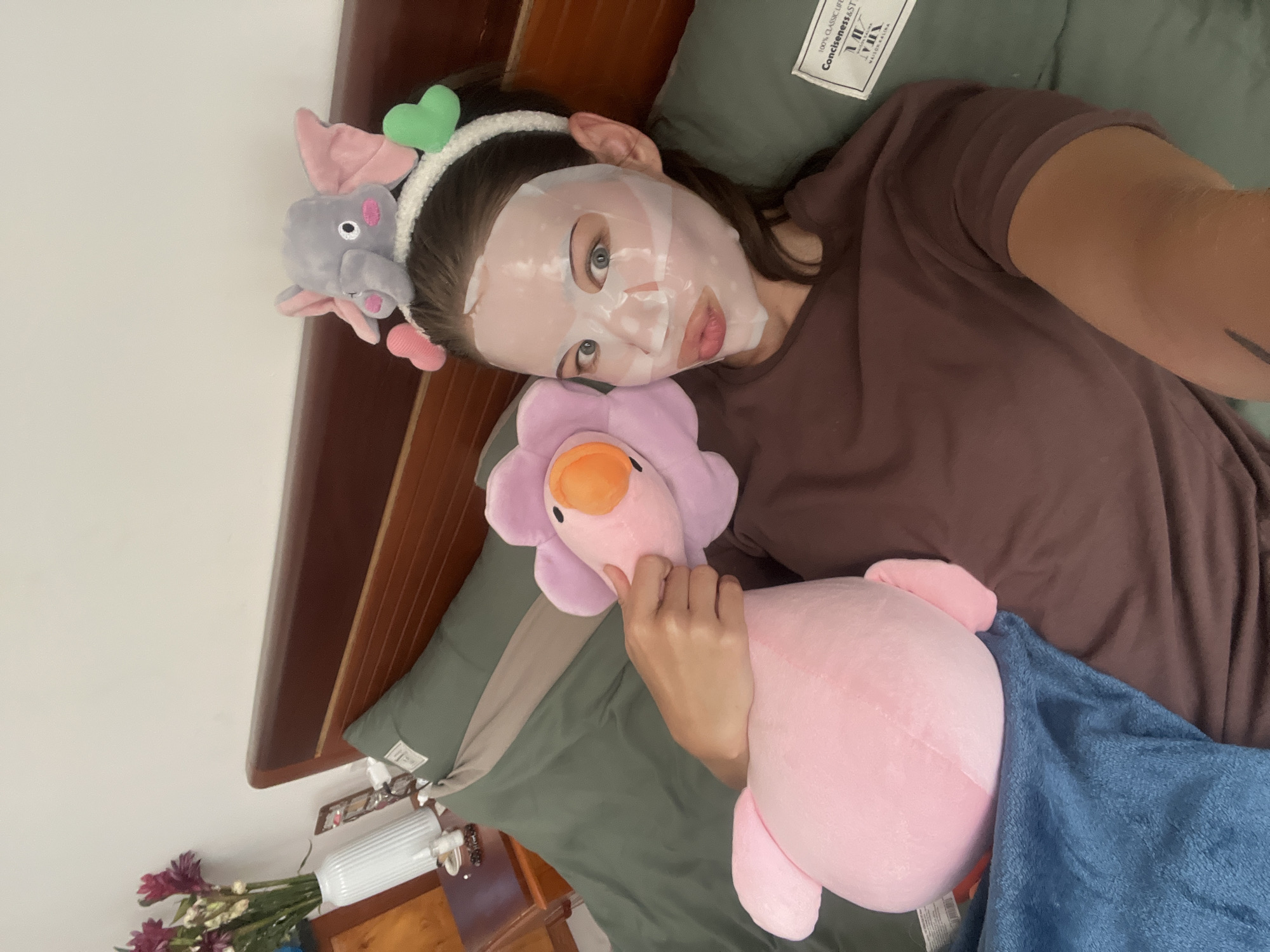 A woman wearing a face mask and a headband with a stuffed elephant and hearts lies on a bed with a stuffed flower and a blue blanket. The bed has green sheets and pillows. A wooden headboard is visible. A bedside table with flowers and other items is partially visible in the lower left corner. The image was likely taken indoors in Nam Nha Trang Ward, Vietnam.
