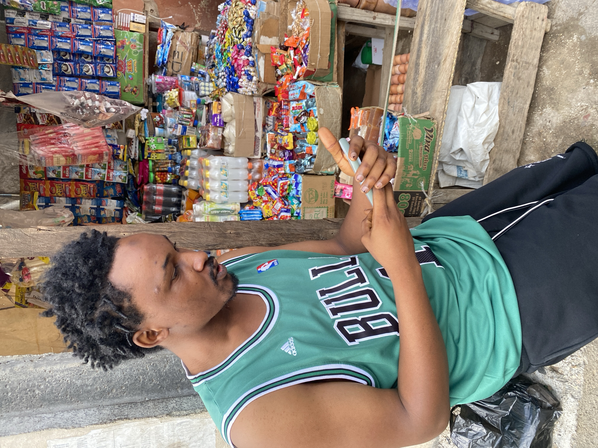 The image depicts a young Black man with dark, curly hair, wearing a green and white sleeveless basketball jersey and black pants. He is seated outdoors, leaning against a textured concrete wall, and looking down at a light blue object he is holding in his hands, which appears to be a phone or similar device. In the background, a small open-air market stall is visible, stocked with a variety of colorful packaged goods, including snacks, drinks, and toiletries. The stall is constructed from weathered wood. The ground is a mix of concrete and dirt. The overall impression is one of a casual scene in a marketplace environment in Jos, Nigeria.