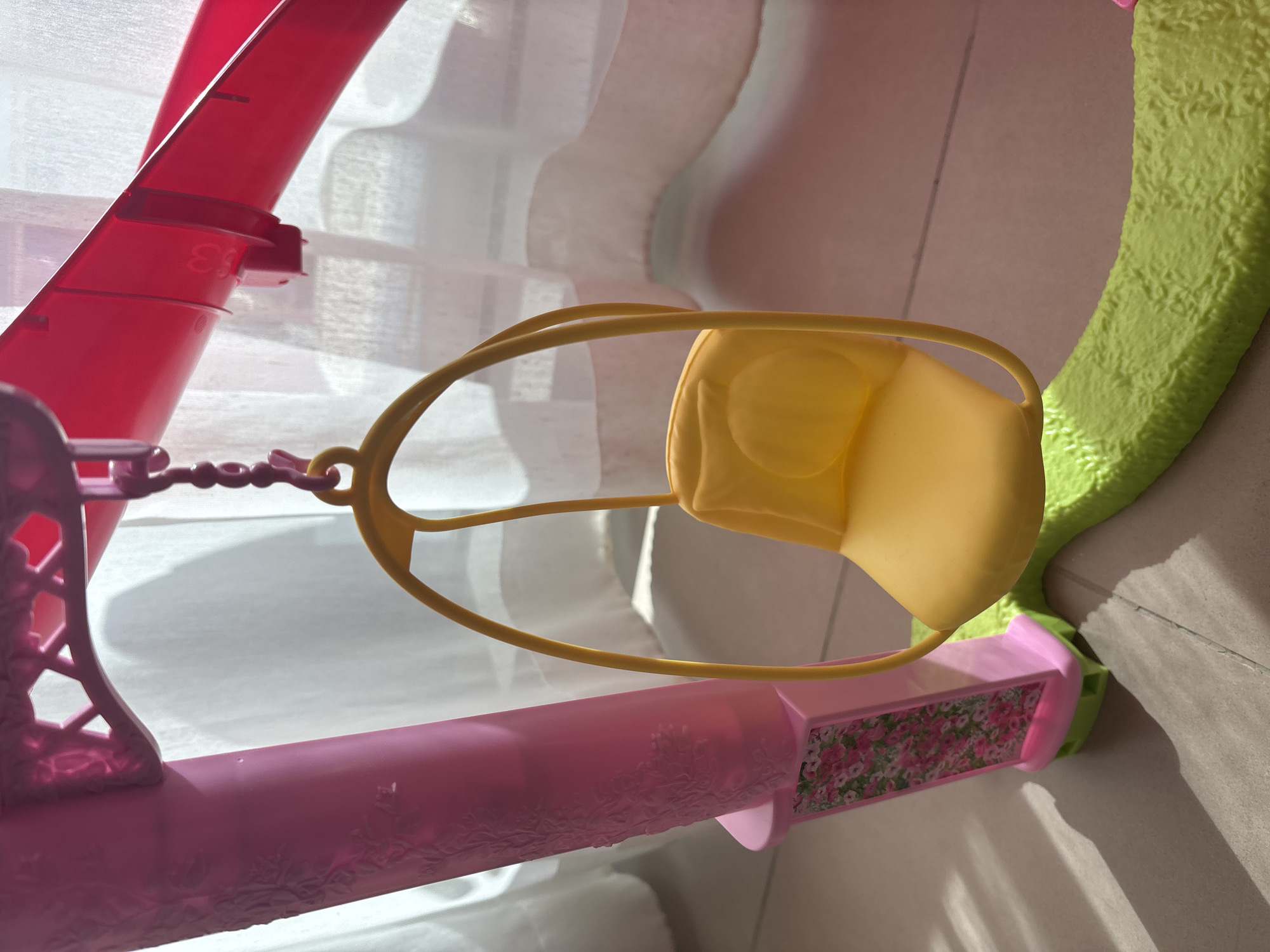 An indoor, close-up, eye-level shot depicts a pink and yellow toy swing set, presumably for dolls. The swing seat is bright yellow and hangs from a pink frame via a pink chain. The frame of the swing set is primarily pink, with a decorative floral pattern on one of the vertical supports. The base of the swing set is a textured lime green. In the background, white curtains are visible, and sunlight casts shadows on the floor tiles. There are no people or animals present in the image, and no discernible activity is taking place. The scene is static and appears to be a still life arrangement of a toy. The context of Dubai, United Arab Emirates, is not visually supported by the image content.