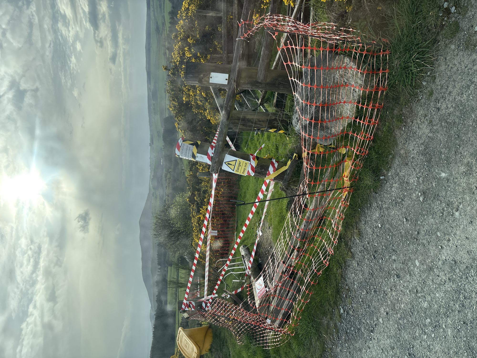 The image shows a rural scene in Shrewsbury, United Kingdom. A damaged wooden gate is heavily barricaded with red and white striped hazard tape and orange plastic netting. Several warning signs are visible, including one with a lightning bolt symbol indicating electrical danger. The gate appears to lead to a grassy field or hillside with yellow flowering bushes. The background features rolling hills and a cloudy sky. The foreground shows a portion of a gravel or dirt road. A yellow bin is partially visible on the lower left. The scene suggests a potential hazard or restricted access area.