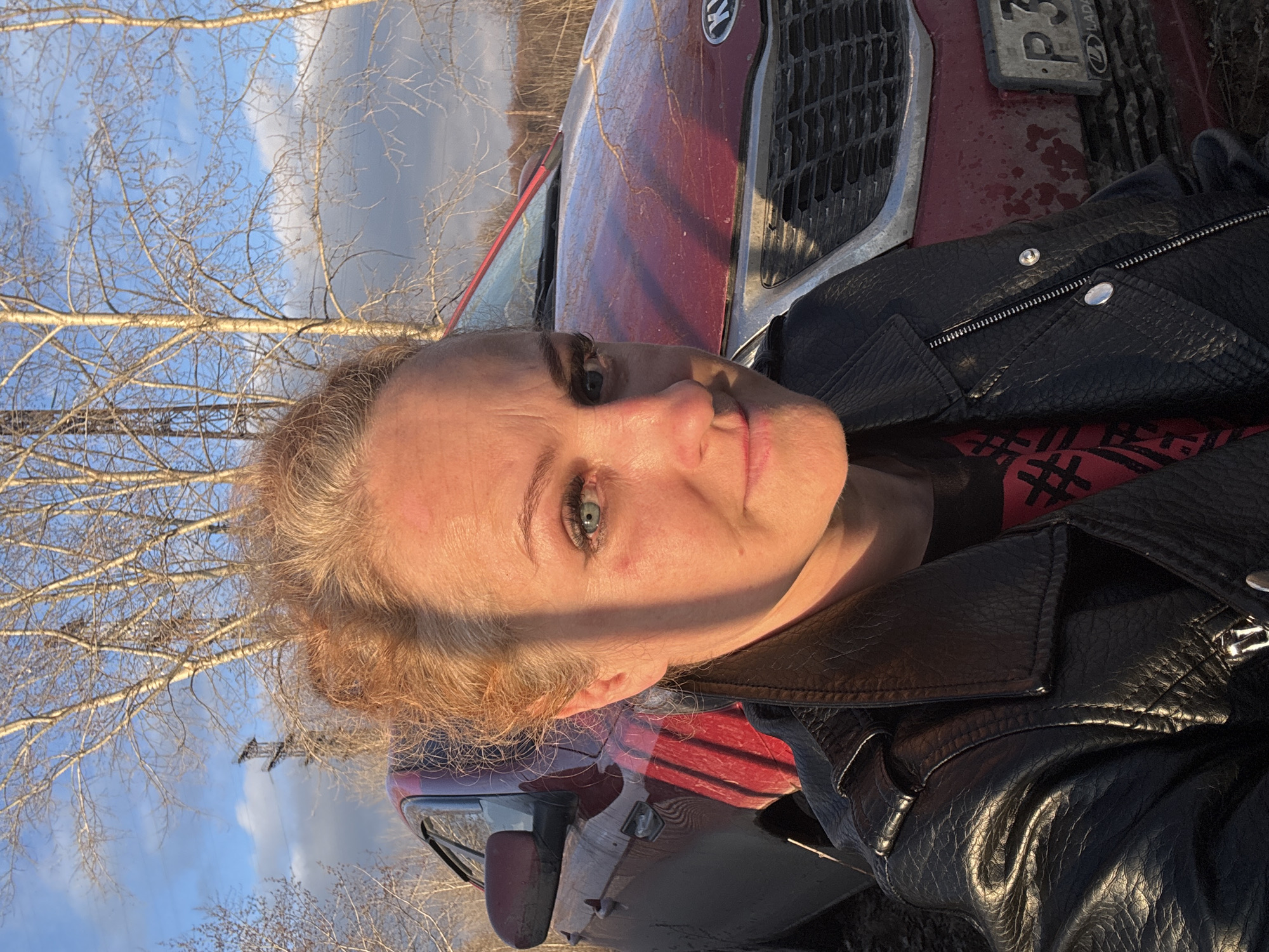 A woman with fair skin, light blonde or light brown hair, and striking blue-green eyes is captured in a close-up outdoor selfie. She wears a black leather jacket over a red shirt with a black pattern or logo. Her expression is neutral as she looks directly at the camera.
She is standing next to or leaning against a red car, identified as a Lada by the visible text on the front grille or below the license plate. A partially visible Russian license plate, featuring Cyrillic characters "P3" and numbers, further confirms the vehicle and location in Russia. The car's hood shows some dirt or dust.
The background features bare tree branches silhouetted against a partly cloudy sky, with patches of clear blue and a large grey cloud. This suggests the season is late autumn, winter, or early spring. Strong, low-angle sunlight illuminates the scene, casting shadows on the woman's face, indicative of either early morning or late afternoon. A faint structure, possibly an electricity pylon, can be seen through the branches. The setting appears to be a rural or semi-rural area in Chaikovsky, Russia.
