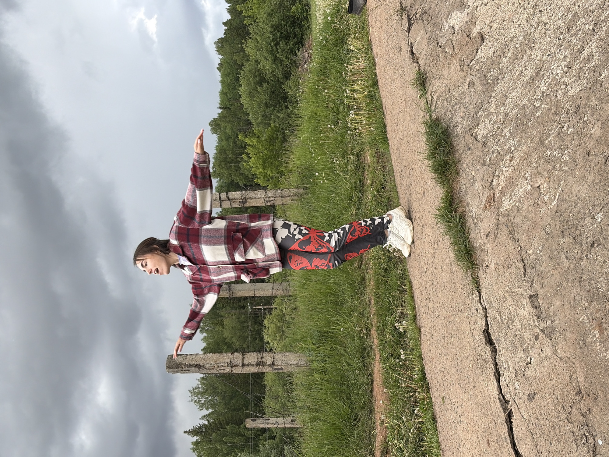 A young person stands with arms outstretched on a concrete edge in Nizhnii Vishur, Russia. The person wears a plaid shirt, patterned pants, and white sneakers. The background features green grass, trees, a cloudy sky, and a concrete fence.