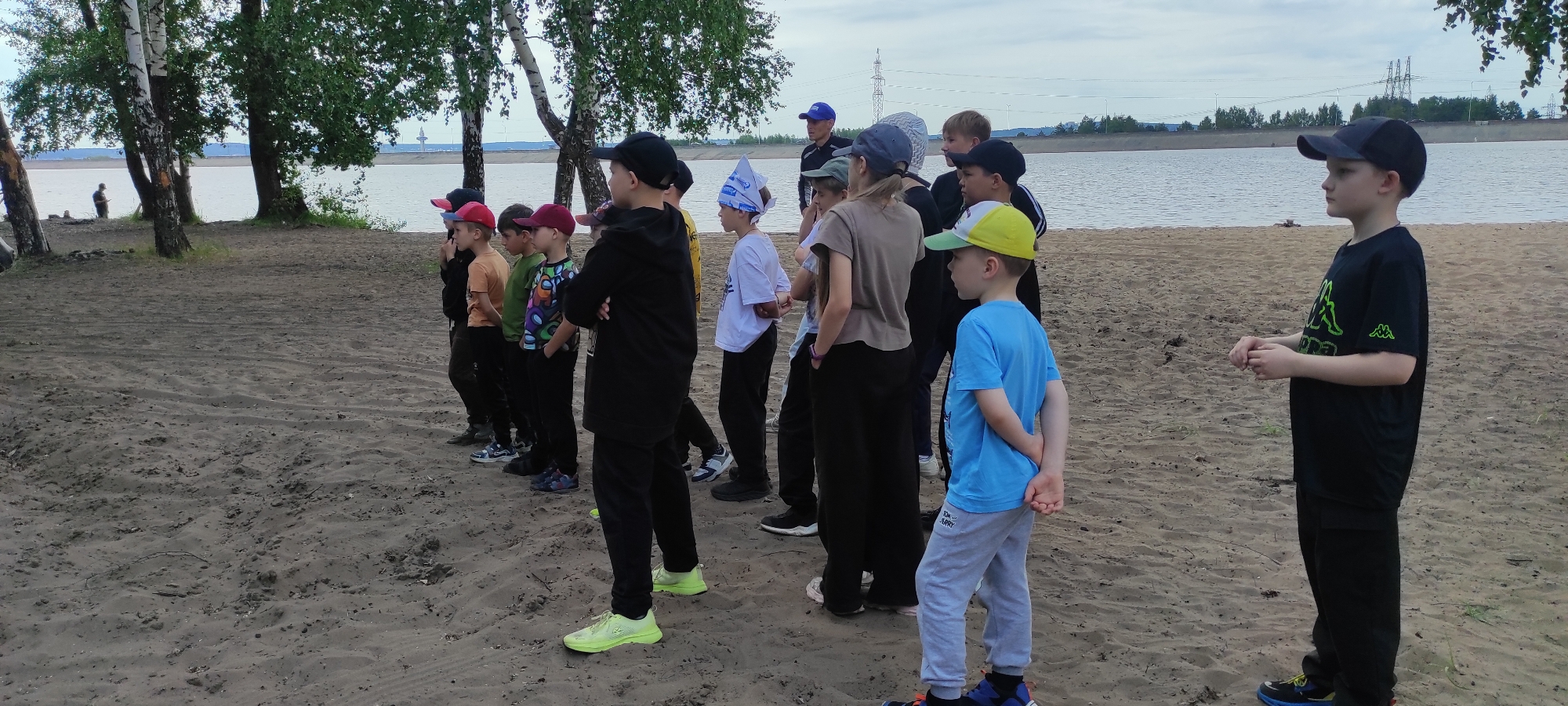A group of children and one adult stand on a sandy shore, facing right. The scene appears to be a beach or riverbank, with a body of water and trees in the background. The sky is overcast, suggesting it might be a cloudy day. The children are dressed in casual, colorful clothing, and most are wearing hats or caps.
The subjects are diverse in age, with the adult in the background wearing a blue cap and dark t-shirt. The children in the foreground are lined up, and some are looking towards the right, while others are looking to the side or down. One child in the middle is wearing a black hoodie and black pants, with their arms crossed. Another child in the front right is wearing a bright blue t-shirt and gray pants with a yellow cap.
There are some trees on the left side of the image, and a person can be seen in the distance on the shore to the far left. The water in the background is calm, and there are some structures visible on the opposite bank, possibly industrial or residential. The overall mood seems relaxed, as if the group is waiting for instructions or an activity to begin.
The logo of Kappa, a sports brand, is visible on the black t-shirt of the boy on the far right. There is also text on the hat of the boy in the middle of the group, but it is not legible. The location is identified as Noviy, Russia.