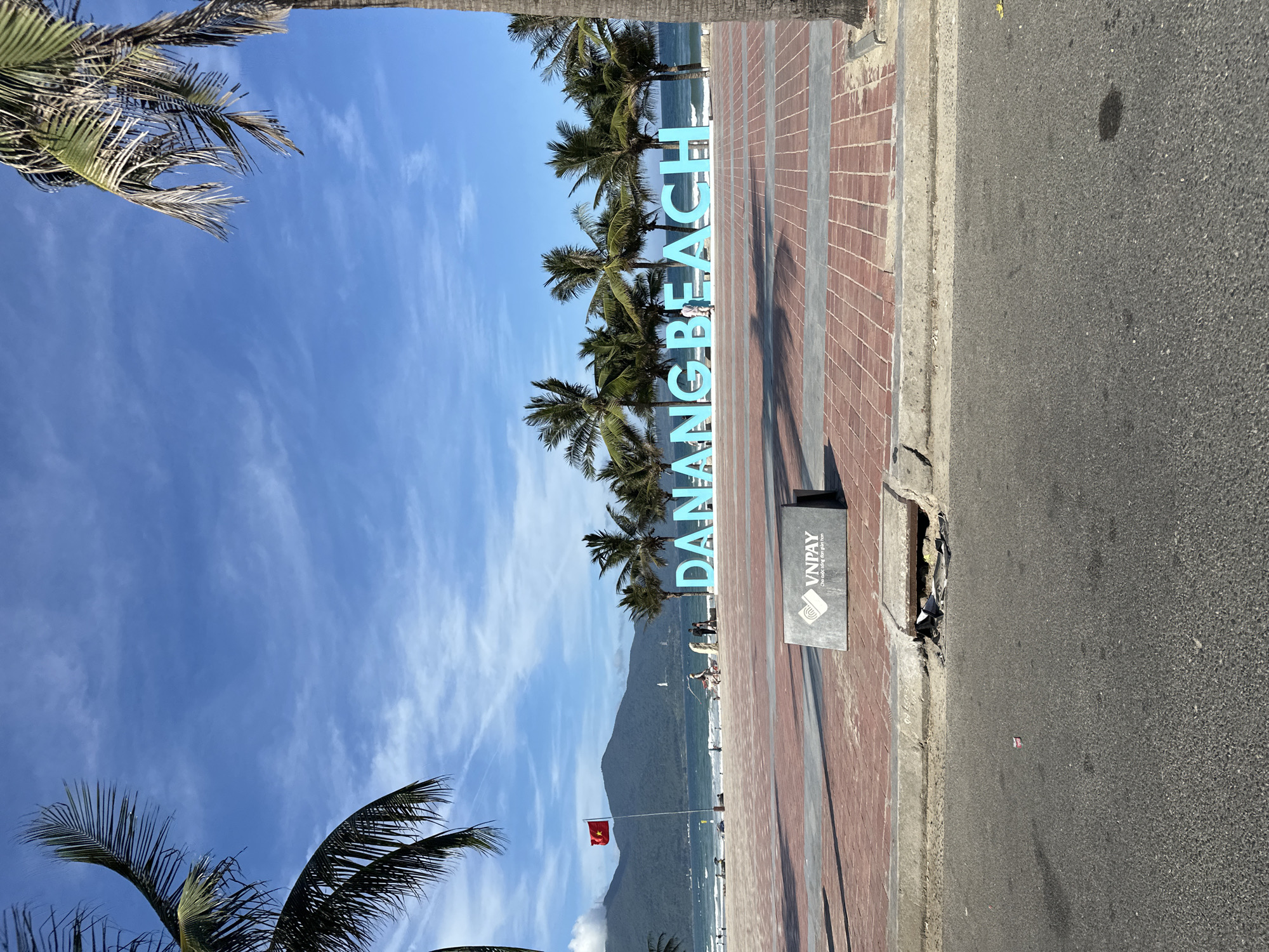 The image depicts the "DA NANG BEACH" sign in Đà Nẵng, Vietnam. The sign is large and blue, standing on a brick-paved sidewalk along the beach. Palm trees are visible behind the sign, lining the beach. In the background, a mountain range is present. A Vietnamese flag flies on a pole closer to the beach. The sky is blue with scattered clouds. A "VNPAY" sign stands near the edge of the sidewalk. A road runs alongside the sidewalk. People are sparsely visible on the beach.