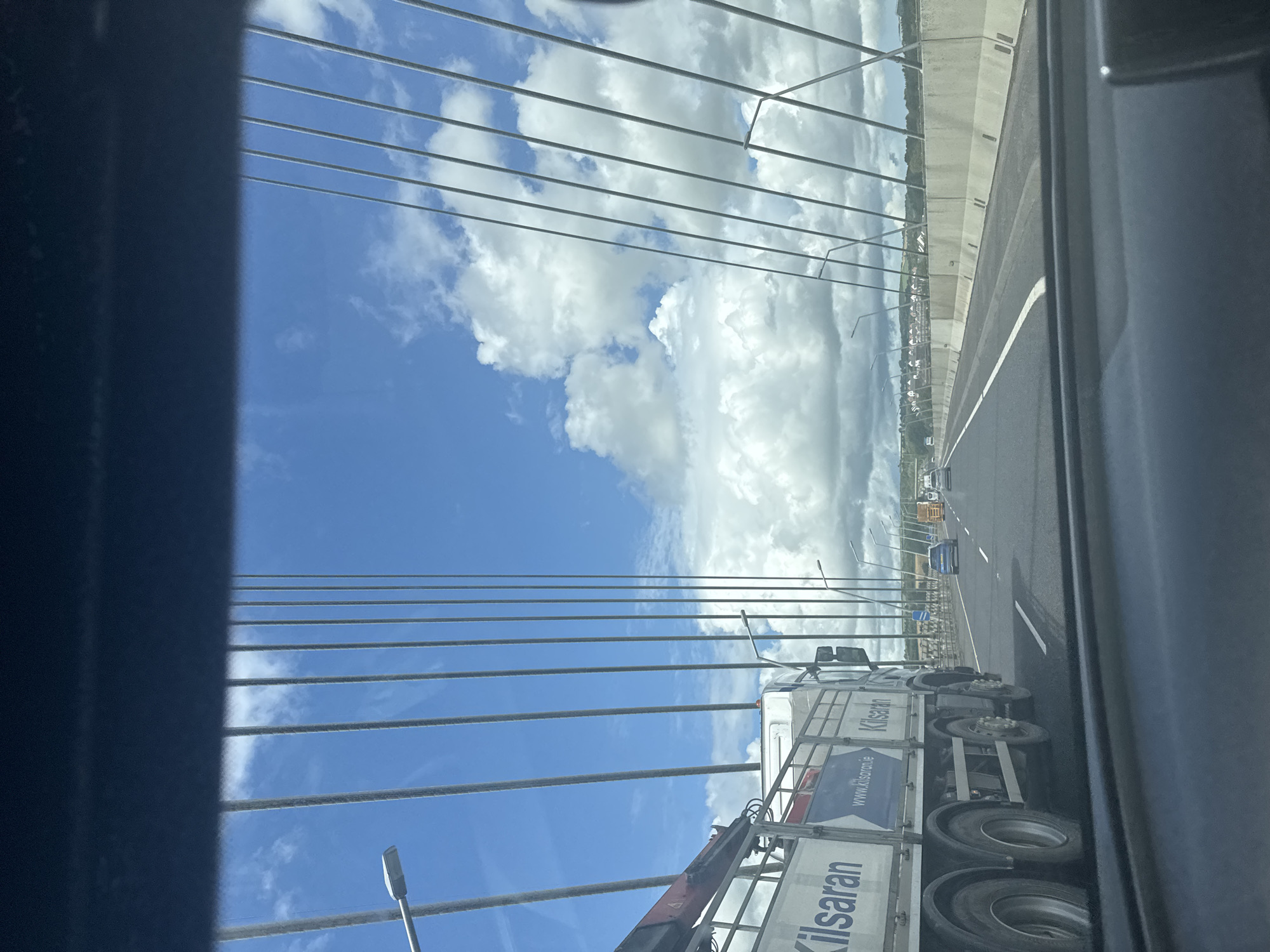 This image, taken from inside a vehicle, shows a view of a bridge and the sky. The perspective is angled, suggesting the photo was taken from a side window. The foreground is dominated by dark, blurred elements, possibly the interior of the vehicle. Stretching across the frame are numerous parallel suspension cables, a defining feature of the bridge's structure.
Beyond the cables, a bright blue sky with fluffy white clouds is visible. The sky takes up a significant portion of the image, creating a sense of openness. A highway runs horizontally across the middle ground, with a white dividing line visible. Several vehicles, including a truck and a car, are driving on the highway. The truck has a white trailer with blue and white text and a logo. The text "Kilsaran" is clearly visible on the side of the trailer, along with "www.kilsaran.ie".
Further in the distance, a treeline and buildings can be seen along the horizon, suggesting a populated area beyond the bridge. Lampposts line the side of the highway. The lighting indicates it is daytime, likely with good weather due to the clear sky and bright clouds. The overall impression is a snapshot of travel on a bridge on a clear day.