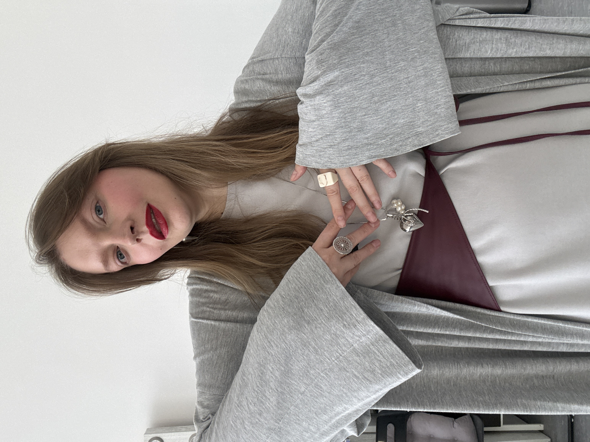 A woman with long brown hair, red lipstick, and fair skin is posing. She is wearing a gray robe-like garment with large, bell-shaped sleeves, under which is a light gray garment with a dark red V-shaped accent piece. She has a white square ring on her right hand and a silver, filigree-style ring on her left hand. A silver necklace with a pendant of a beetle-like insect with pearls is positioned in front of her chest. The background is a plain white wall. The bottom of the frame shows a glimpse of a black object and a gray cushion or bag. There is no indication that the location is Izhevsk, Russia.
