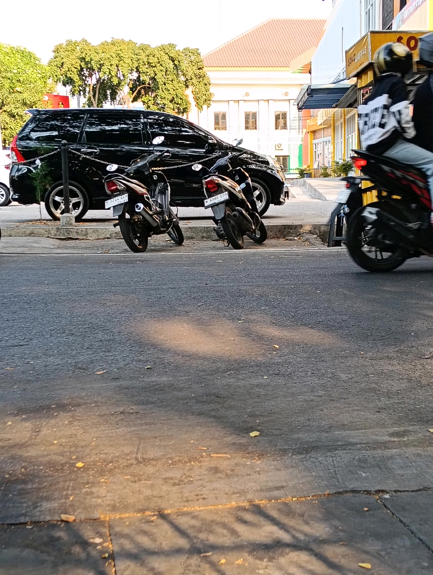 In the city of Karanganyar, Indonesia, on a sunlit street, a black minivan is parked alongside a curb. Two motorcycles are parked behind the minivan, one black and one red and black. A third motorcycle, also black, is in motion to the right of the frame, its rider wearing a helmet. The street is paved, with a raised concrete sidewalk. Trees with dense green foliage line the background, behind a building with a red tiled roof. The building also features white walls and multiple windows.