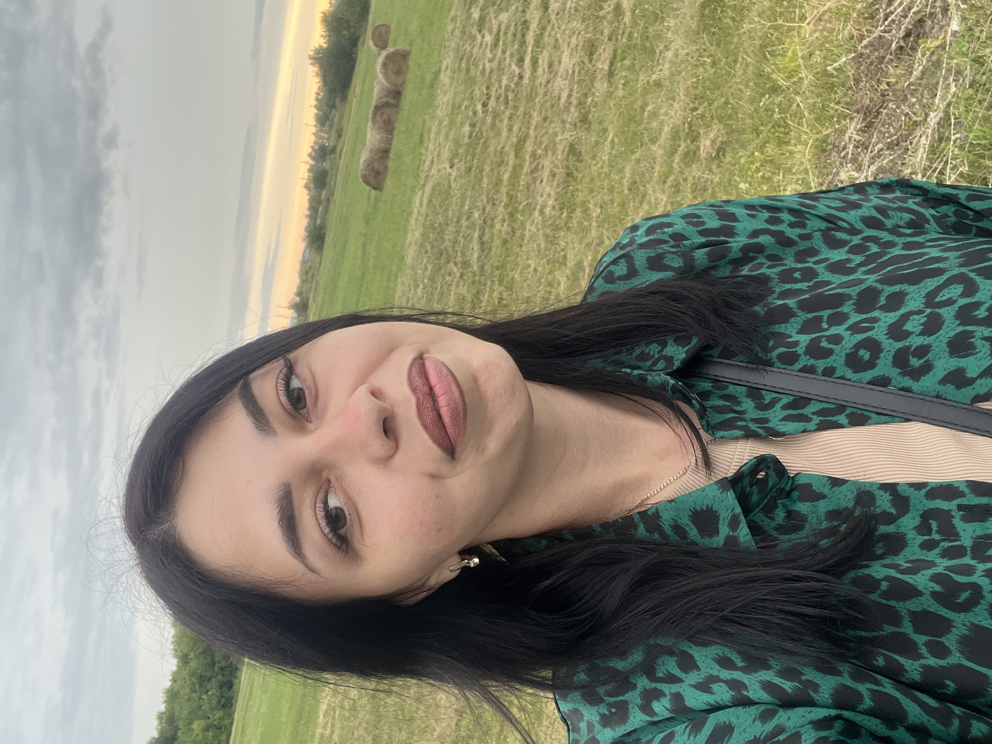A medium shot captures a woman with long, dark hair and medium skin tone from the chest up, facing the camera directly. She is outdoors in a grassy field with scattered hay bales in the background, under a cloudy sky with a hint of sunset colors on the horizon. The woman wears a green and black leopard-print button-down shirt over a beige tank top. She has on light-colored earrings and a delicate necklace. Her makeup includes brown lipstick and mascara. To her right, a dark strap, possibly from a bag, is visible across her chest. The setting appears to be a rural area, potentially on the outskirts of Istobnoye, Russia, given the natural landscape.
