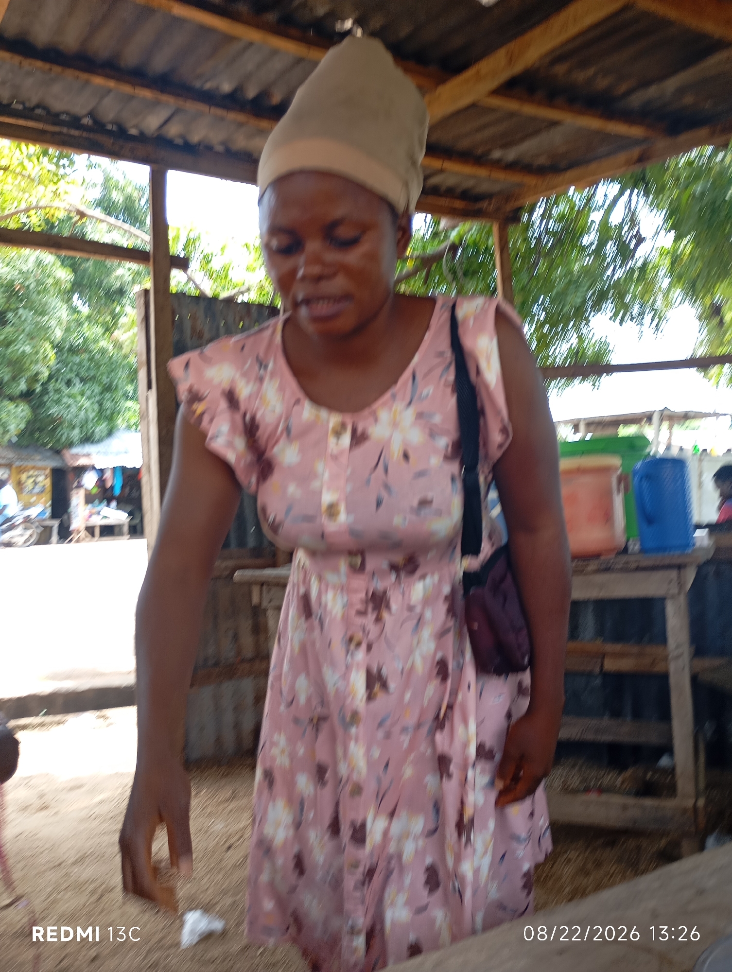 The image shows a woman standing outdoors under a corrugated metal roof supported by wooden beams. She is wearing a light pink dress with a floral pattern and a beige head covering. A dark-colored bag is slung over her right shoulder. Her left hand is extended downwards, and she appears to be interacting with something on the sandy ground. Behind her, a wooden table holds two large containers, one orange and one blue. In the background, trees and other structures are visible, suggesting a market or roadside stall setting. The overall lighting indicates it is daytime. The timestamp "08/22/2026 13:26" and "REDMI 13C" are present in the lower corners of the image, suggesting the image was taken with a Redmi 13C phone on that date and time. The scene appears to be in Shendam, Nigeria, as indicated by the provided context.