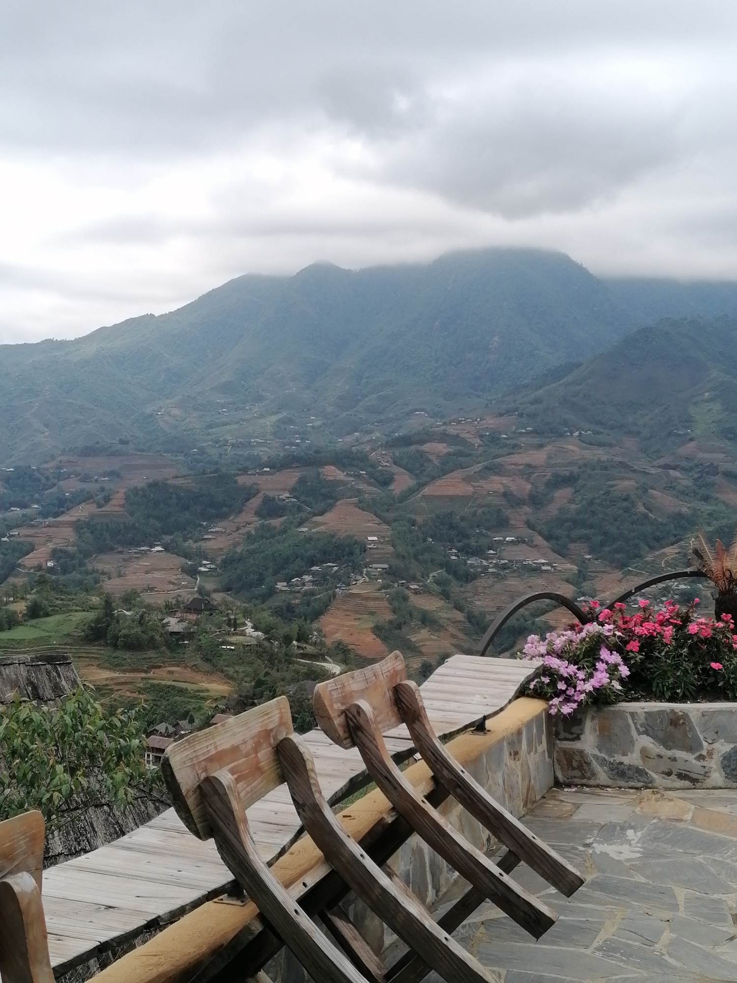 The image depicts a mountainous landscape in Sa Pa Ward, Vietnam, viewed from an elevated platform. In the foreground are wooden chairs positioned along the edge of the platform. A flowerbed containing pink and purple flowers is located nearby. The middle ground reveals terraced fields and scattered villages across the undulating terrain. The background features a mountain range shrouded in clouds. The weather appears overcast.