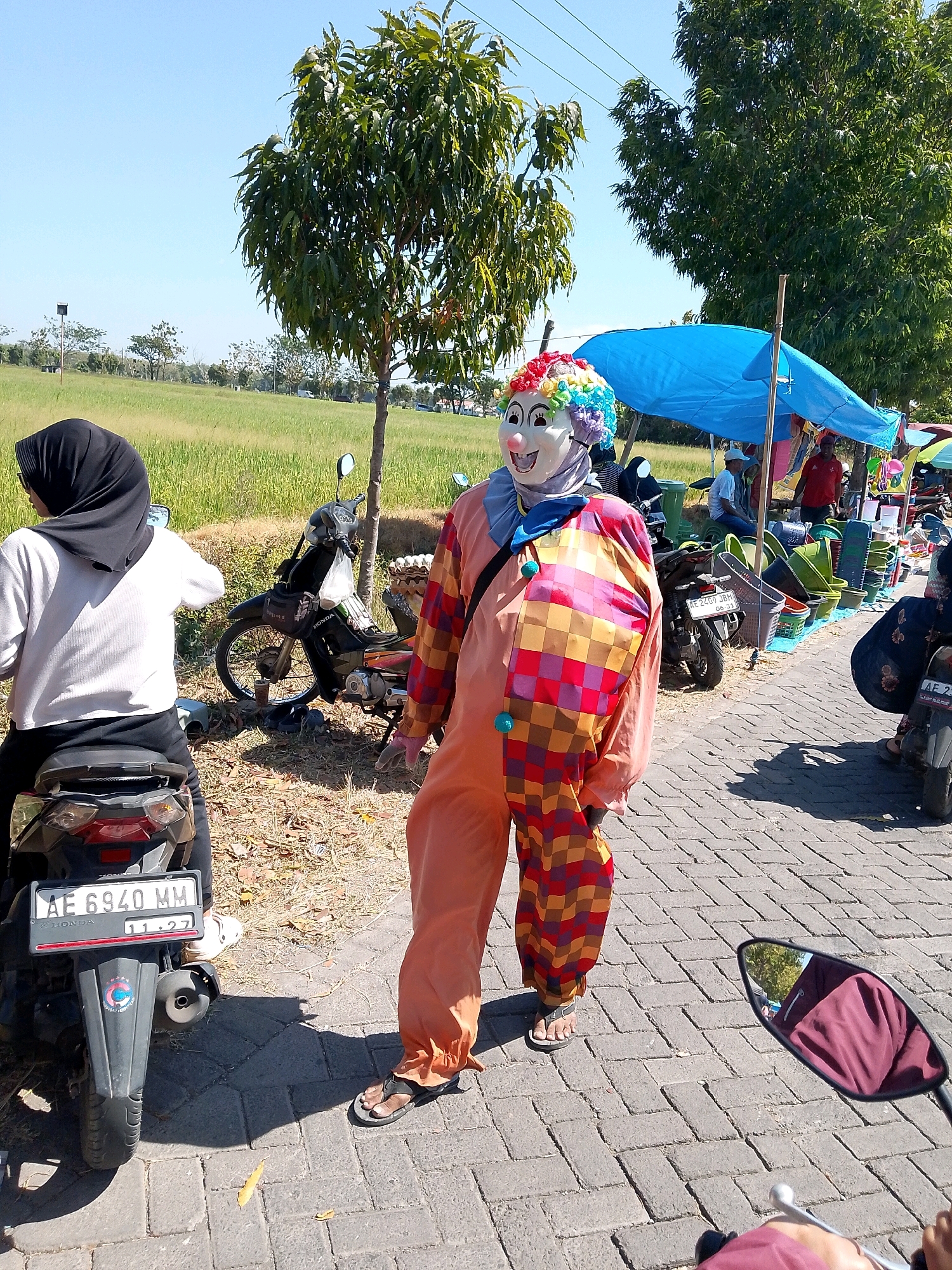 A person in a clown costume stands on a brick pathway in Geneng, Indonesia, on a sunny day. The costume is orange with a checkered pattern of red, yellow, and purple squares on the front. The clown's mask is white with large painted eyes and a red smile, topped with a colorful wig. To the left of the clown is a black motorcycle with a license plate reading "AE 6940 MM". Behind the motorcycle, a person wearing a black hijab and a white long-sleeved shirt is seated on another motorcycle. In the background, there is a field of green grass, trees, and a blue tarped canopy covering a market stall. The market stall displays various plastic containers. Other motorcycles and people are visible around the stall. The sky is clear and blue.