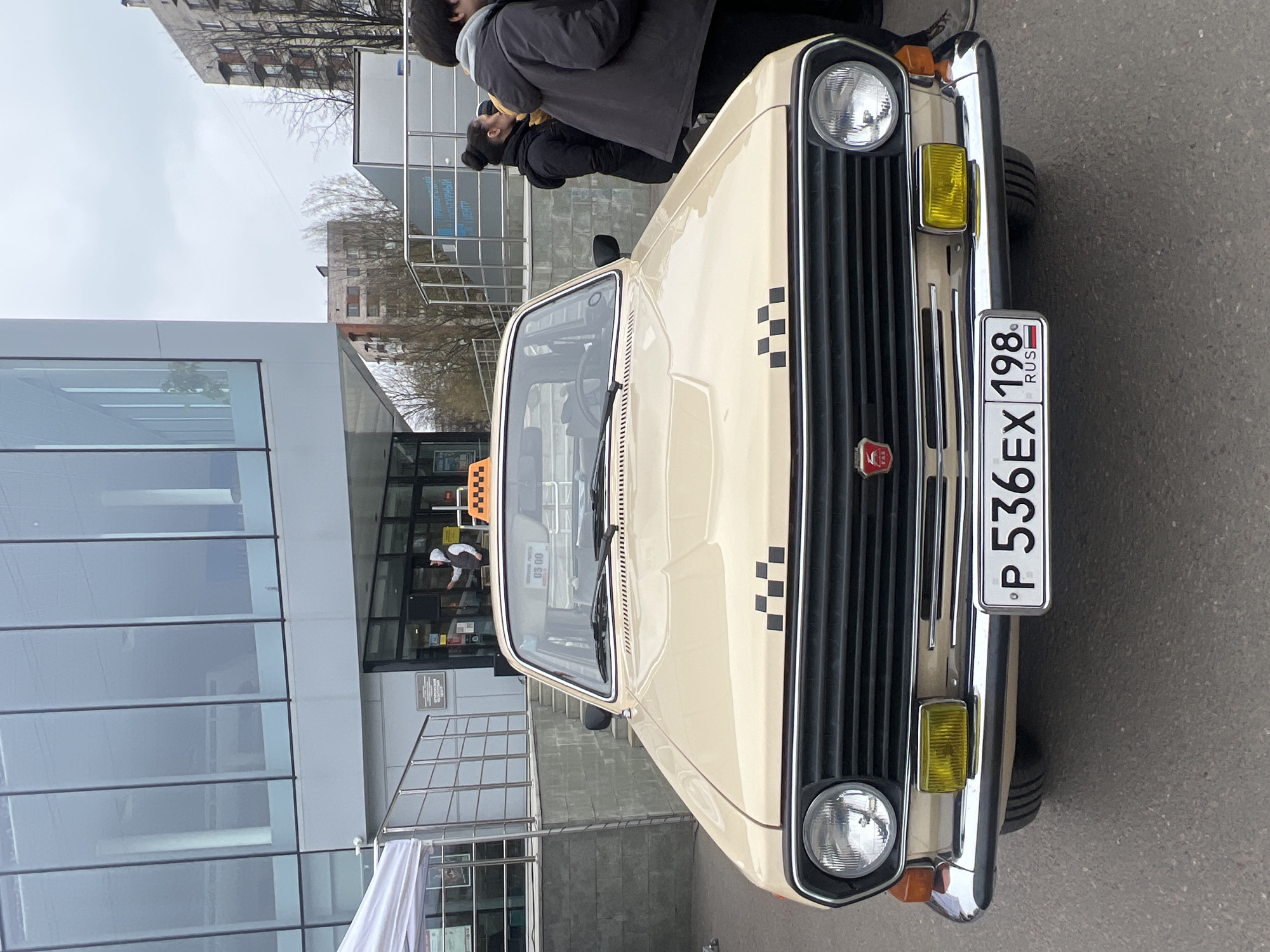 A light yellow taxi is parked on an asphalt street in front of a modern glass-walled building. The front of the car is in the foreground, with a Russian license plate reading "P 536 EX 198 RUS". The taxi's hood has black checkered taxi markings. People are standing in the background near the building. The location is the city of Saint Petersburg, Russia.