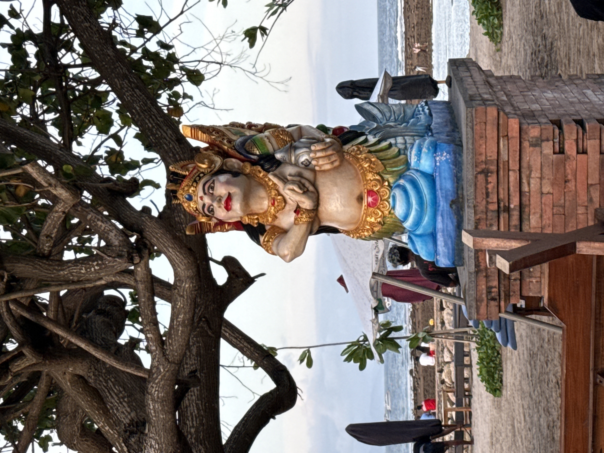 A partially obscured view features a colorful, ornate statue positioned near a beach in Denpasar, Indonesia. The statue, portraying a figure with golden adornments and blue garments, is positioned on a brick structure. The beach is visible in the background, with people, vegetation, and structures scattered along the shoreline. A prominent tree with thick branches and foliage stands on the left, partially framing the statue. The sky is overcast, and the overall environment suggests a coastal area in a populated tourist destination.