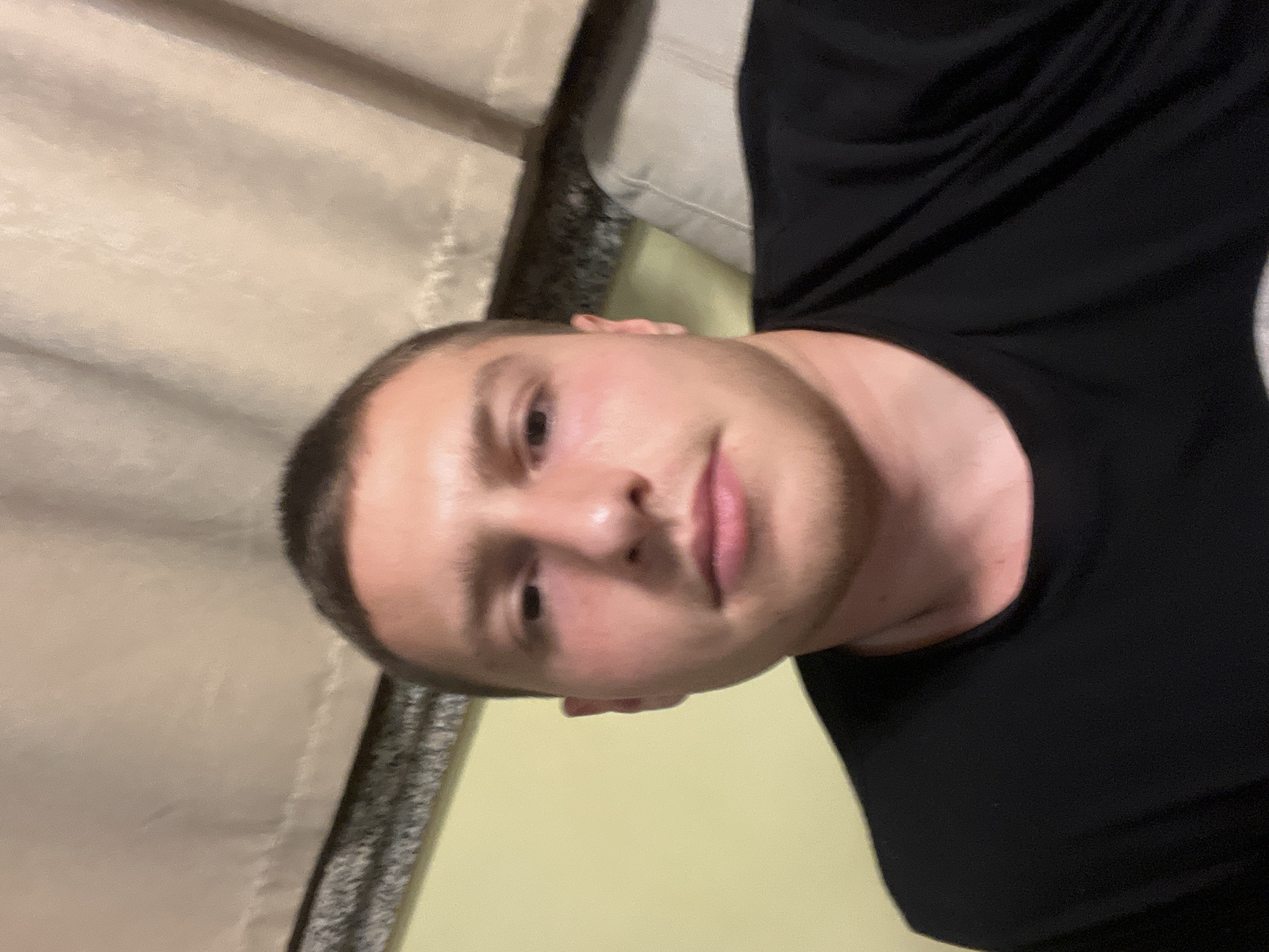 A man with short, dark hair is wearing a black t-shirt. The background includes a curtain and part of what appears to be a sofa. No location information is provided.
