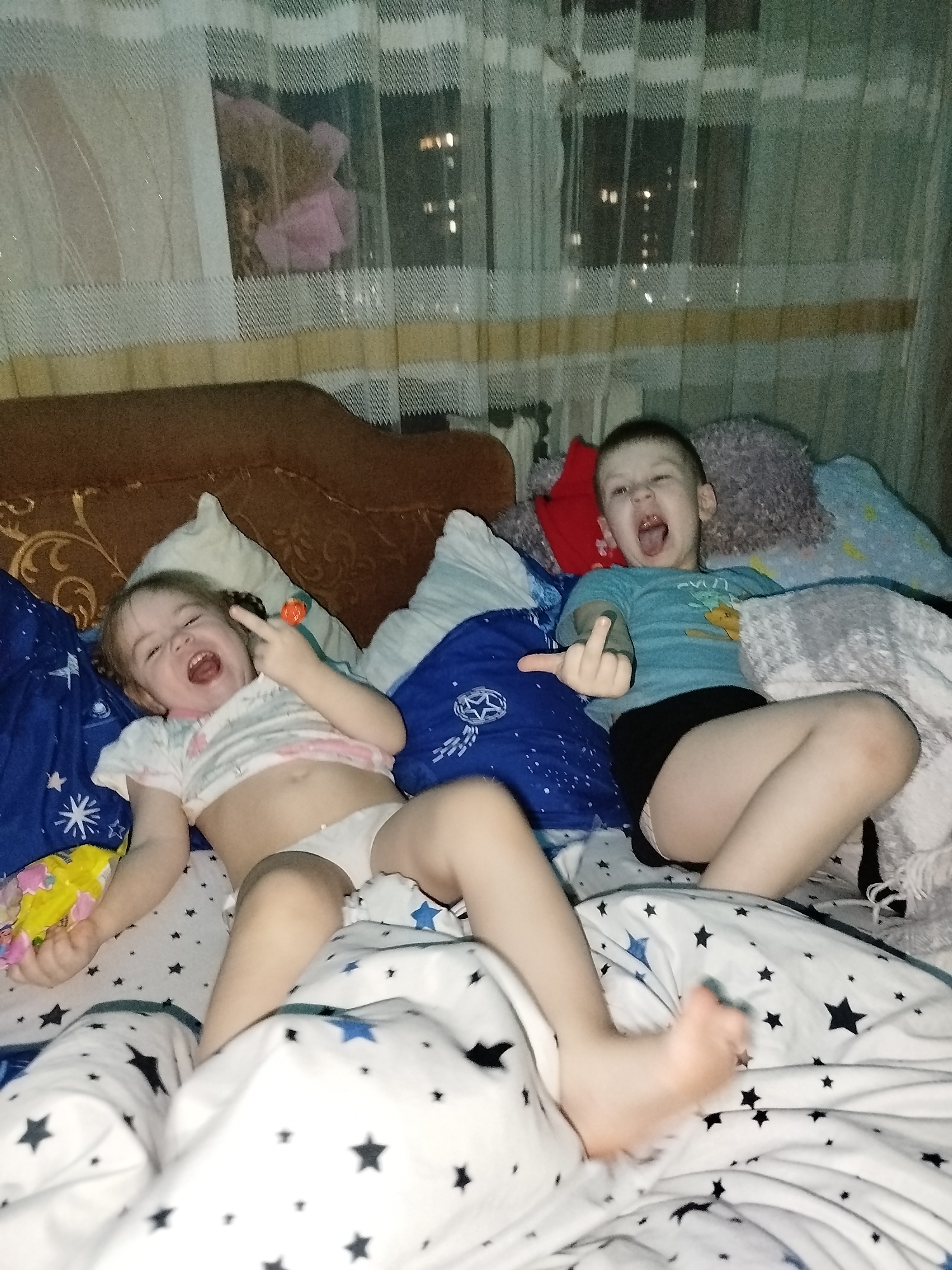Two young children, a girl and a boy, are lying on a bed with a star-patterned comforter. The girl is wearing a white shirt and underwear, and she is holding a toy in her left hand while extending her right middle finger. The boy is wearing a blue t-shirt and black shorts, and he is also extending his right middle finger. Both children have their mouths open in what appears to be shouting or laughter. The background includes pillows, a brown headboard, and a window with translucent curtains. Through the window, lights suggest a nighttime urban scene, indicating the location is in a city; it is assumed to be Chaikovsky, Russia.
