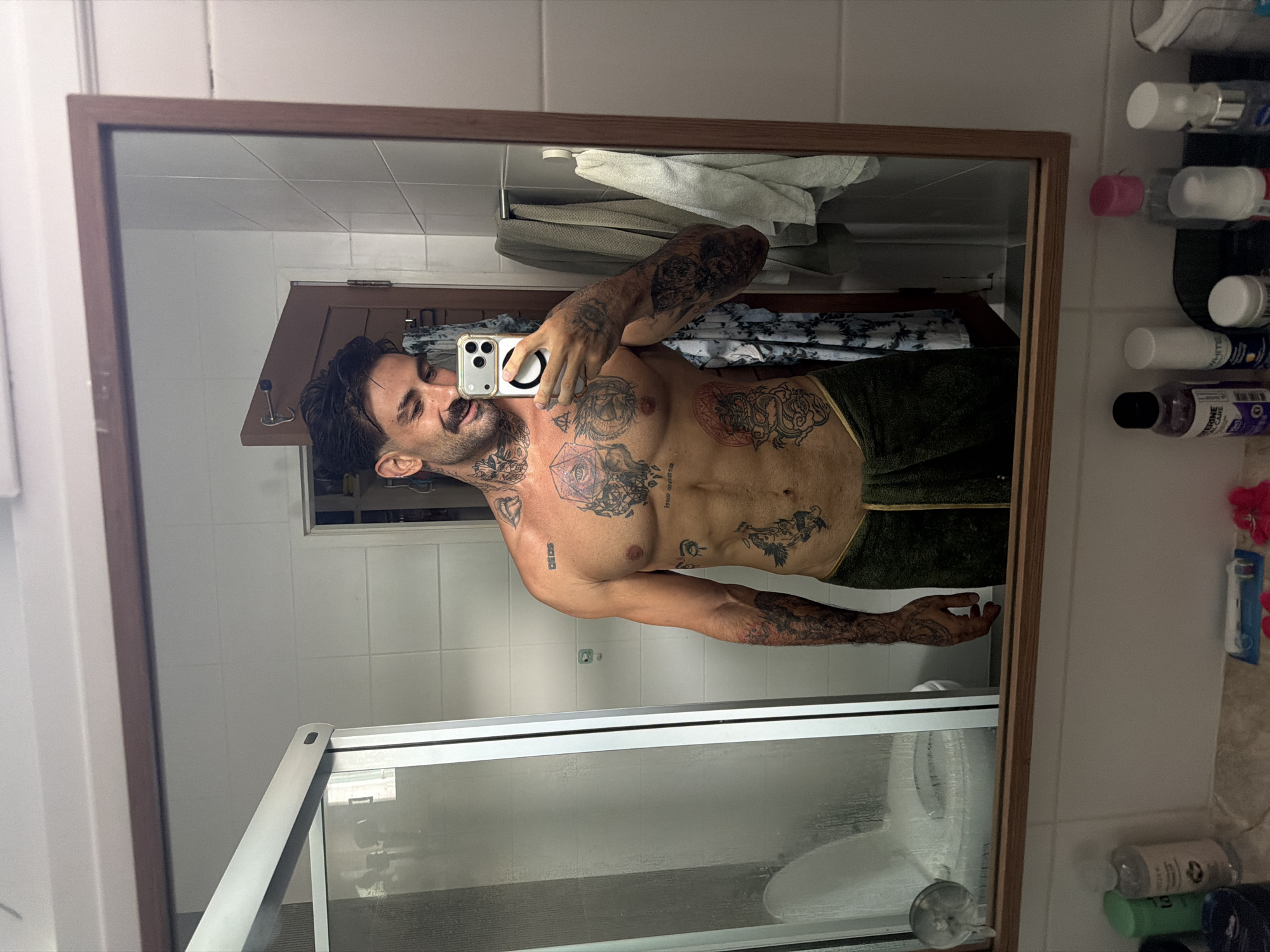 A shirtless man stands in a bathroom, wearing a dark green towel wrapped around his waist. He is holding a smartphone, taking a selfie in the mirror. He is fair-skinned with dark hair, and has extensive tattoos on his chest, abdomen, arms, and neck. The bathroom has white tiled walls. In the background, there are white towels and a shower curtain. To the right of the mirror are various toiletry products. A shower with a sliding door is visible below the mirror. The image was taken in the city of Chalong, Thailand.