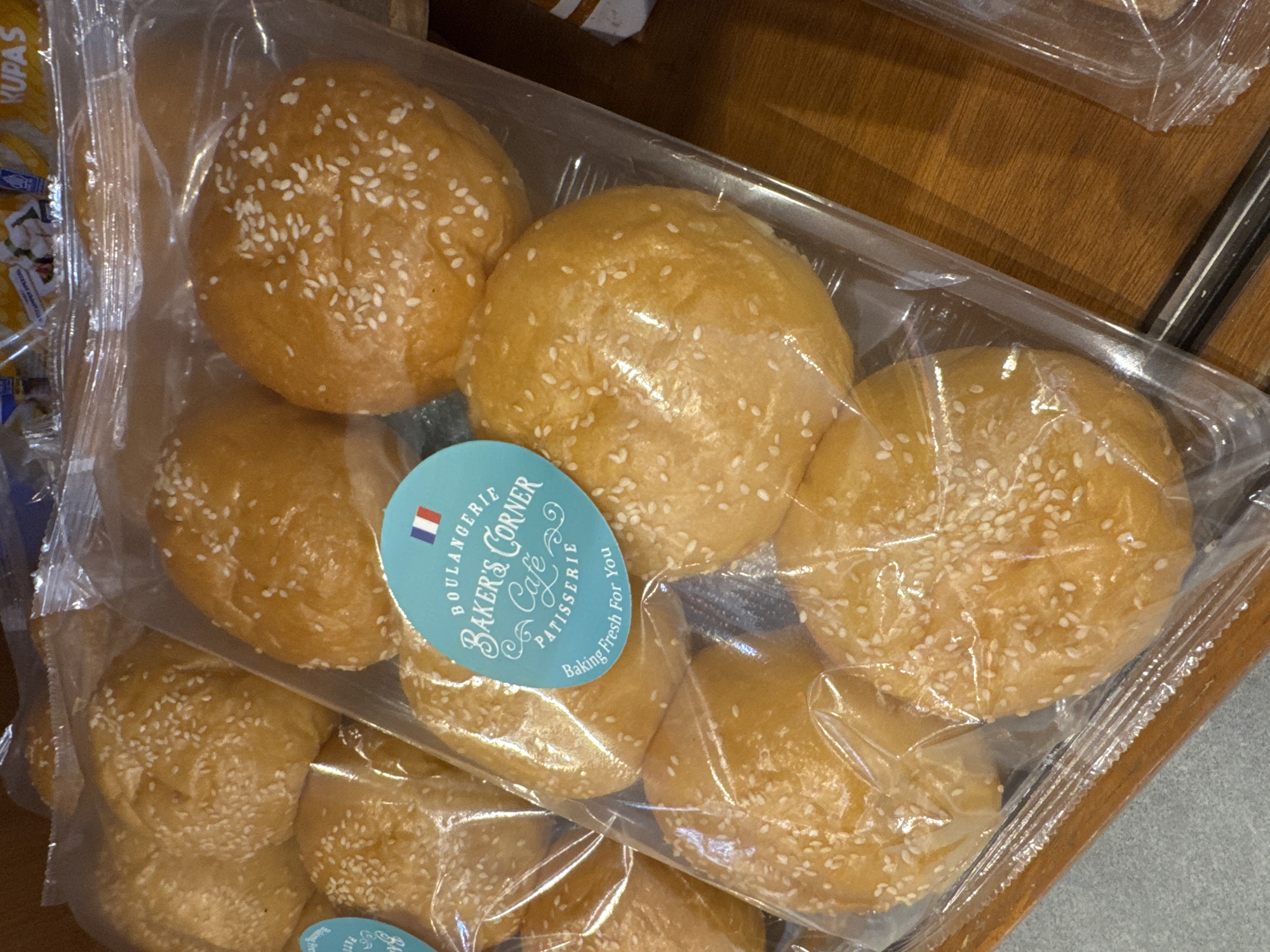 A package of sesame seed buns from Boulangerie Baker's Corner Cafe Patisserie are in a clear plastic package. The label shows the french flag and says, "Baking Fresh for You." The package is on a wooden surface. Kuta Selatan, Indonesia, is not discernible from the image.