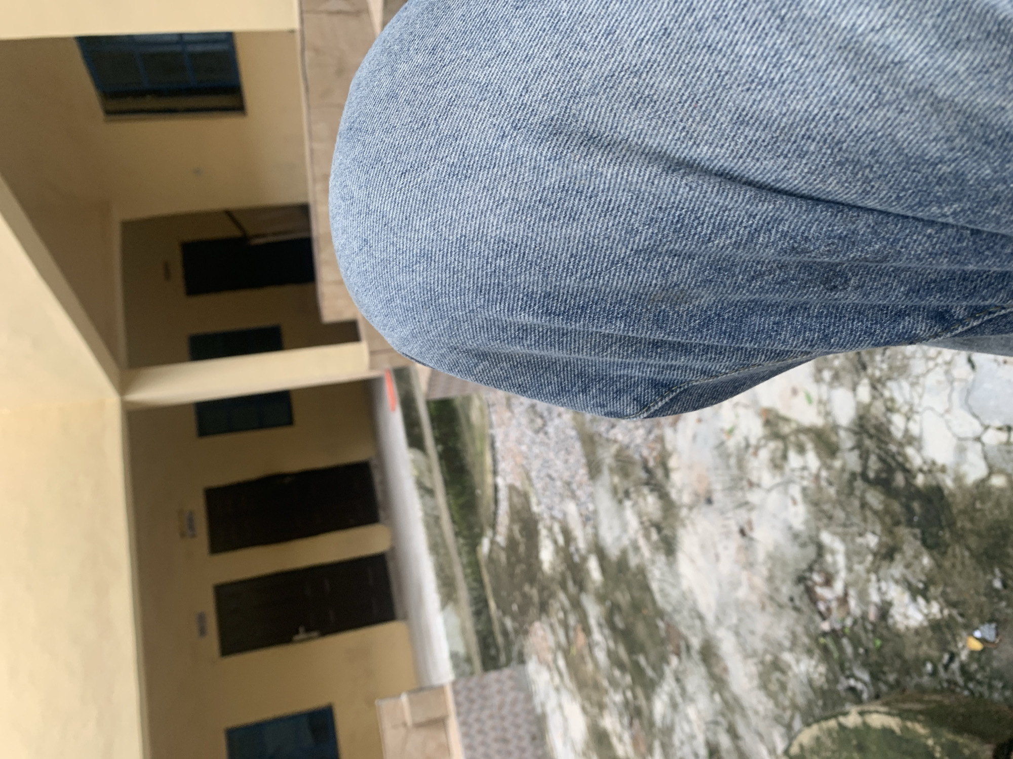 The image shows a close-up of light blue denim jeans in the foreground, suggesting a person's legs are in the frame. The background is a blurred view of an outdoor environment, possibly a courtyard or walkway. To the left, there's a section of a building with light yellow walls. It appears to have multiple doorways, some dark and rectangular, suggesting rooms or offices. Above some of these doorways, there are smaller rectangular windows with blue frames. The ground is visible in the lower right, showing a wet, mottled surface that looks like concrete or stone, possibly after rain. The lighting suggests it could be daytime, but the overall blurriness of the background makes it difficult to ascertain specific details about the time of day or weather conditions beyond the apparent wetness of the ground. The image seems to be taken from a slightly elevated or angled perspective. No text is clearly visible in the image. The context of Jalingo, Nigeria, places this scene in a specific geographical location, but without further visual cues, it's difficult to infer more specific details about the setting within Jalingo itself.