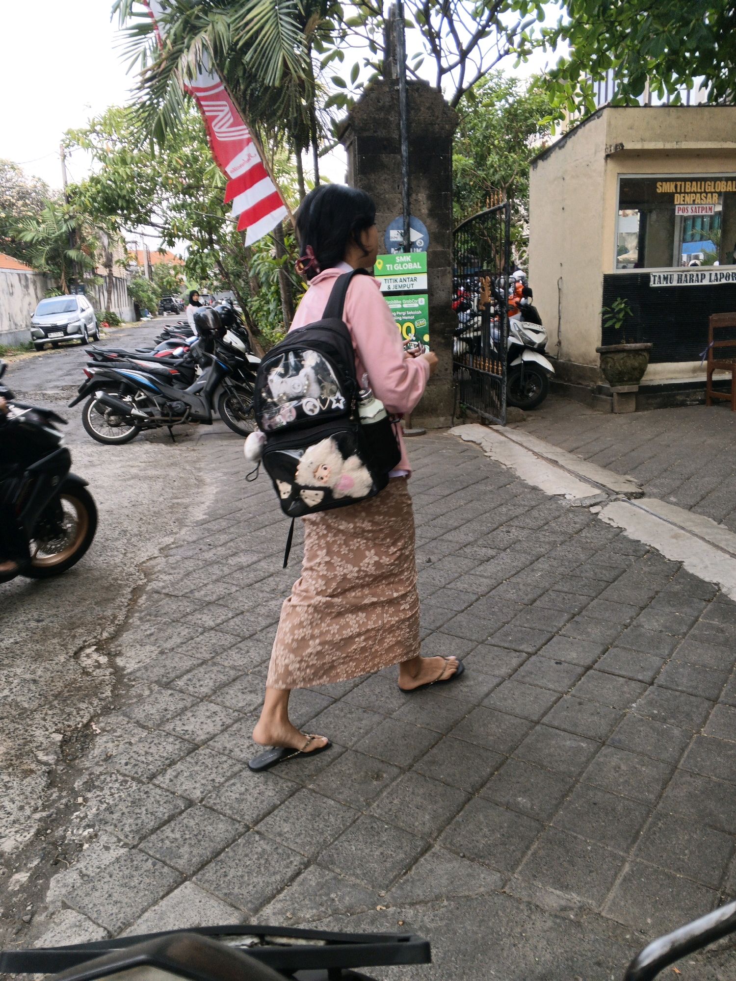 A woman wearing a pink long-sleeved shirt and a long floral skirt walks on a paved sidewalk in Denpasar, Indonesia. She carries a black backpack adorned with plush toys and a water bottle. In the background, motorcycles are parked along the street, and a building with signage indicating "SMK TI BALIGLOBAL DENPASAR" and "POS SATPAM" is visible. A red and white flag hangs from a tree overhead. The environment appears to be a typical street scene in Denpasar, with buildings, vehicles, and pedestrian pathways.